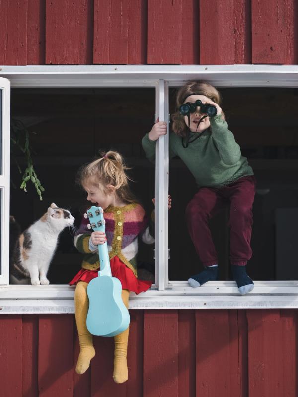 To barn sitter i vinduskarmen, den ene holder en gitar og klapper en katt, mens den andre bruker en kikkert til å se ut.