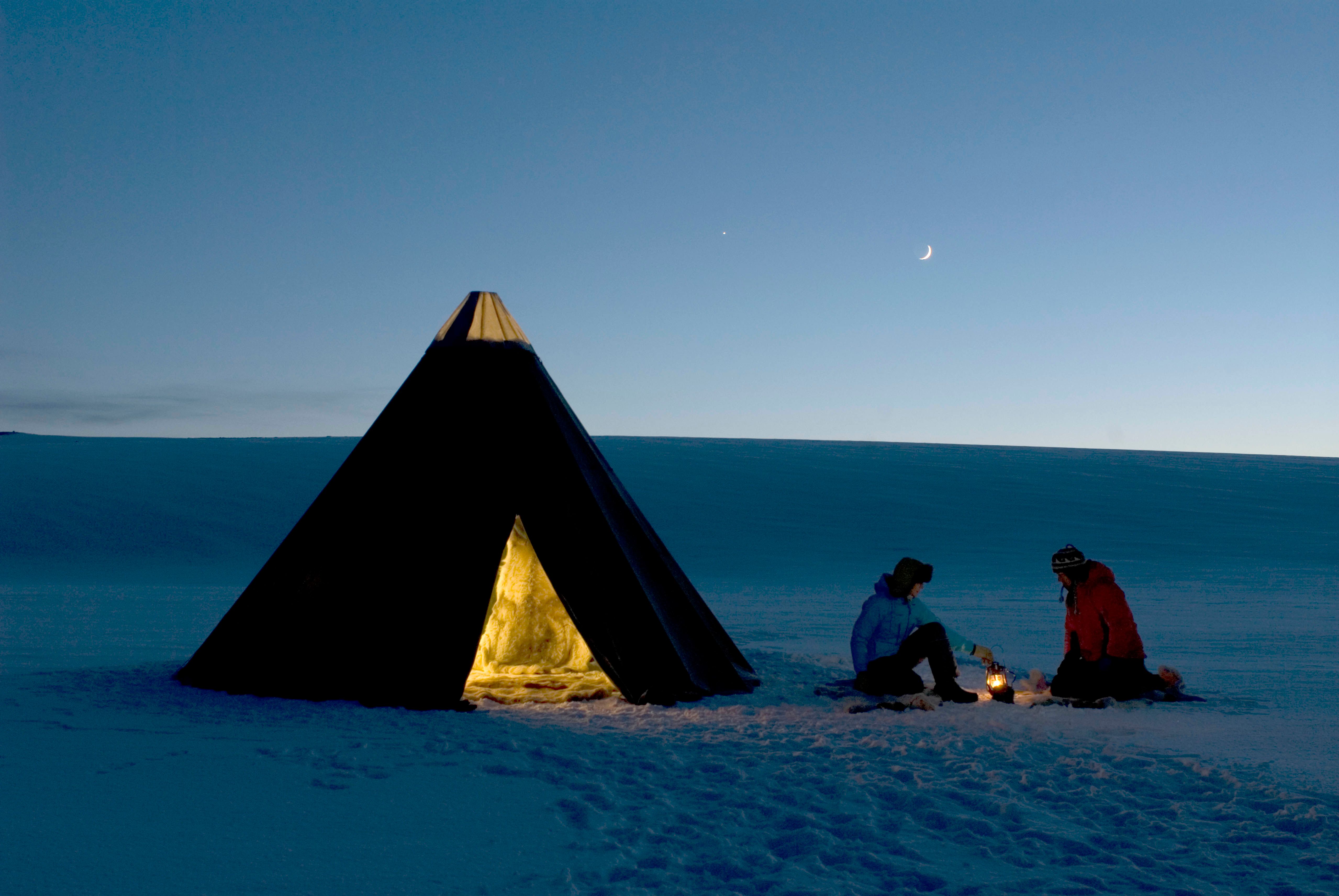 Two people around a fire next to a traditional Sami tent, known as a Lavvo
