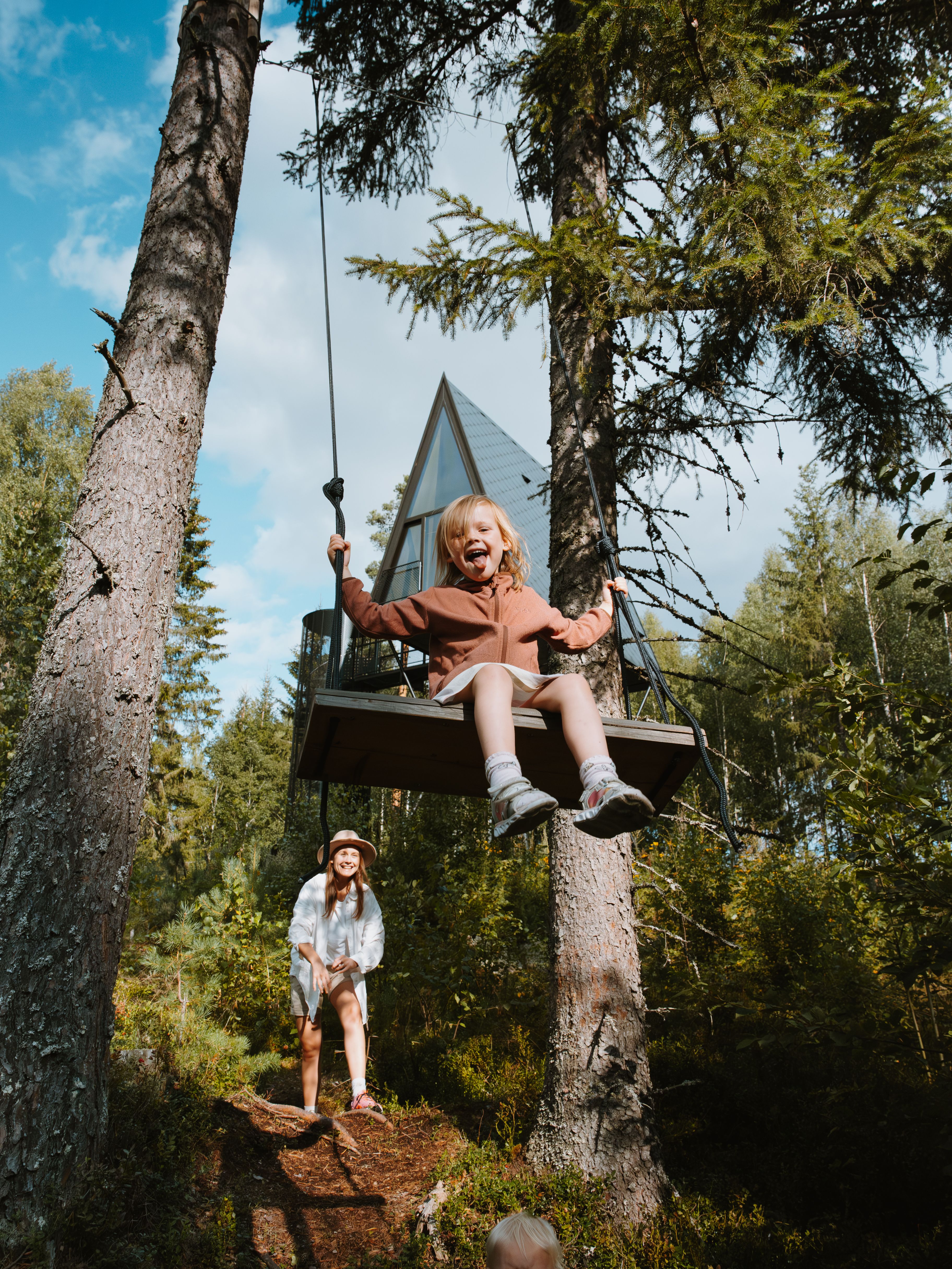 A girl is smiling on a swing in front of a treetop cabin