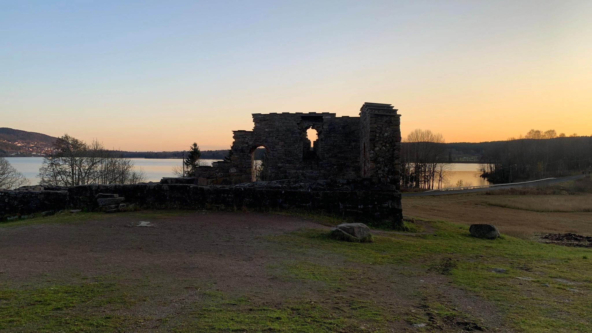 Ruins of a stone structure overlooking a peaceful lake at sunset, surrounded by grasslands and distant hills.