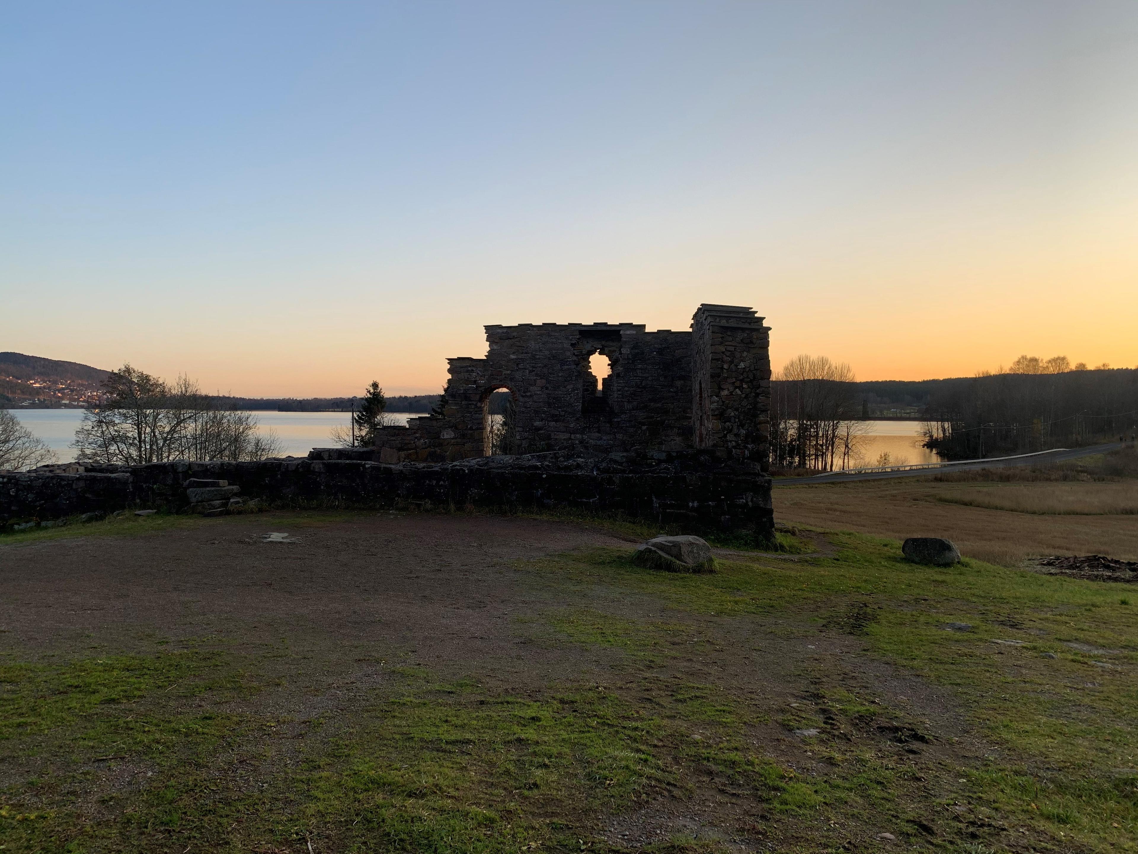 Ruins of a stone structure overlooking a peaceful lake at sunset, surrounded by grasslands and distant hills.