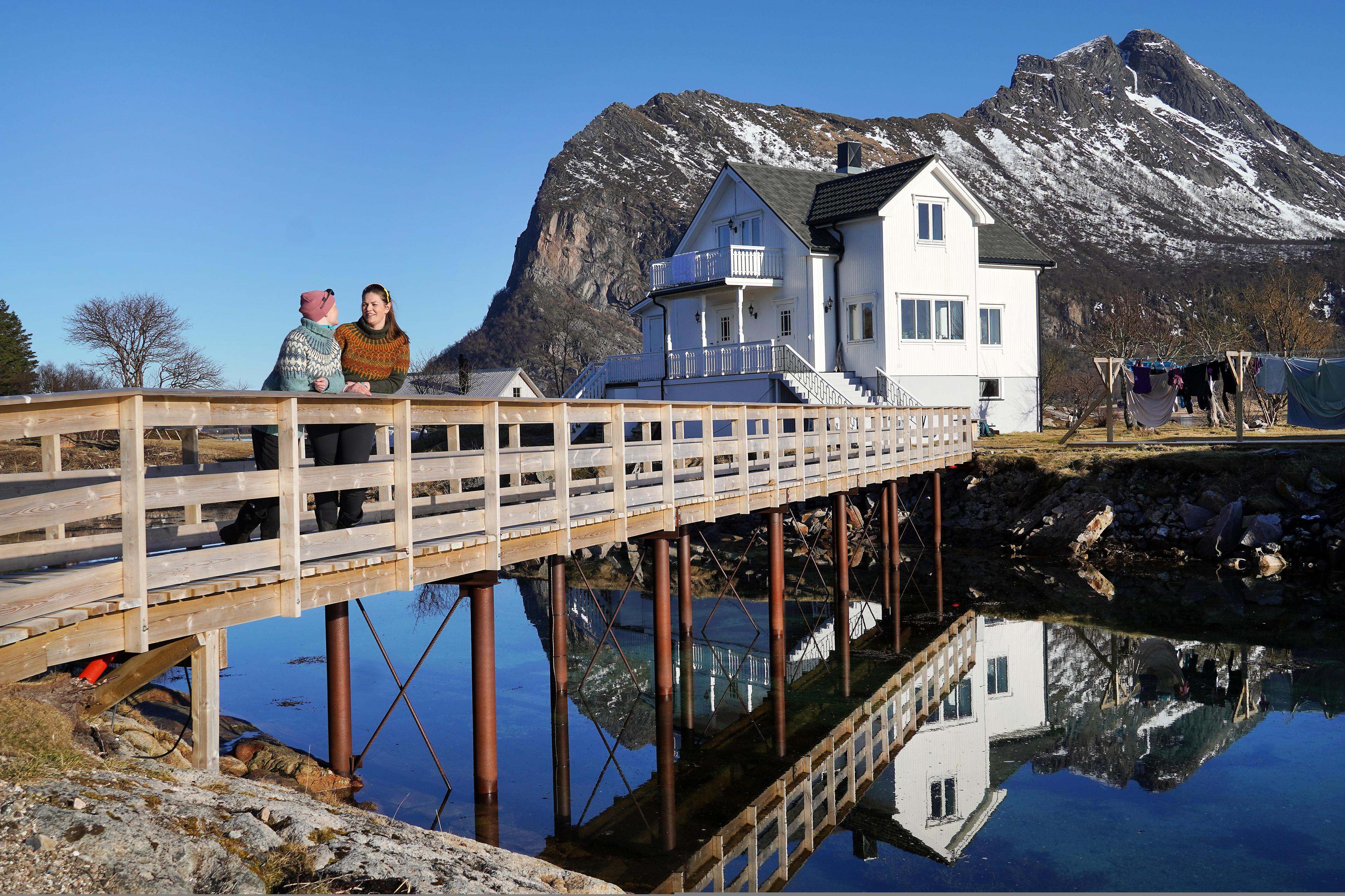 Two women on a bridge in front of a white house and mountains.