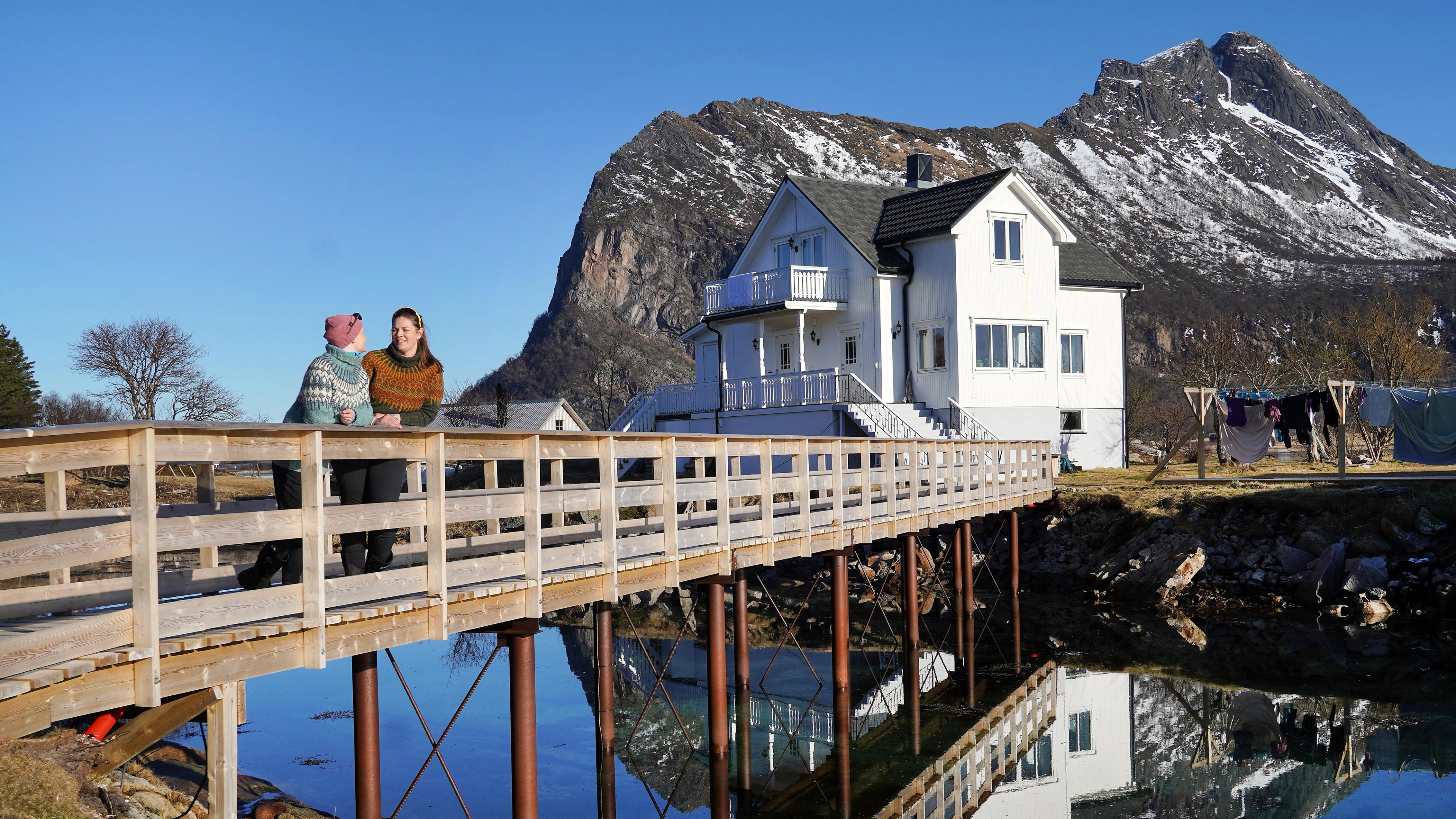 Two women on a bridge in front of a white house and mountains.