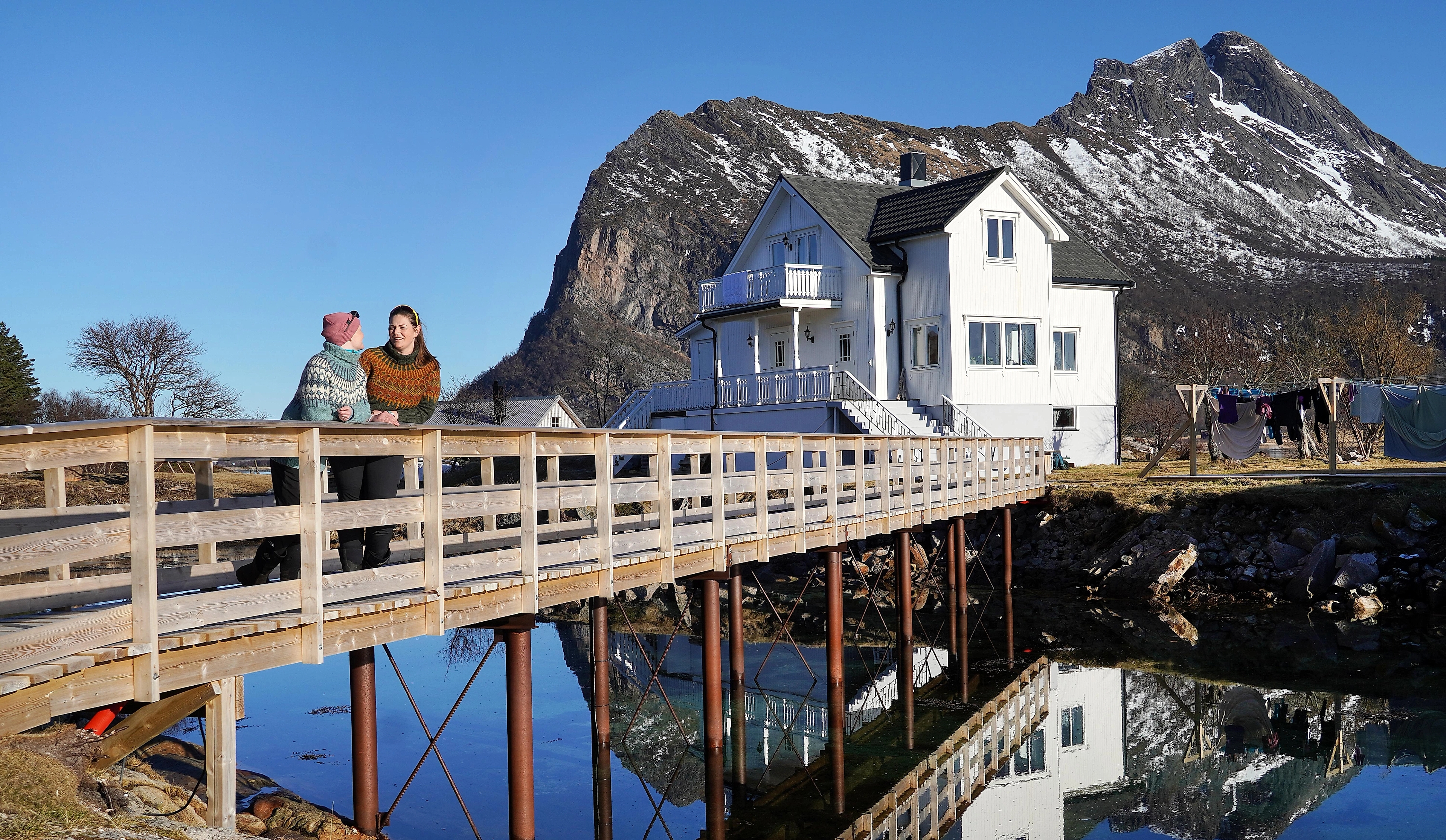 Two women on a bridge in front of a white house and mountains.
