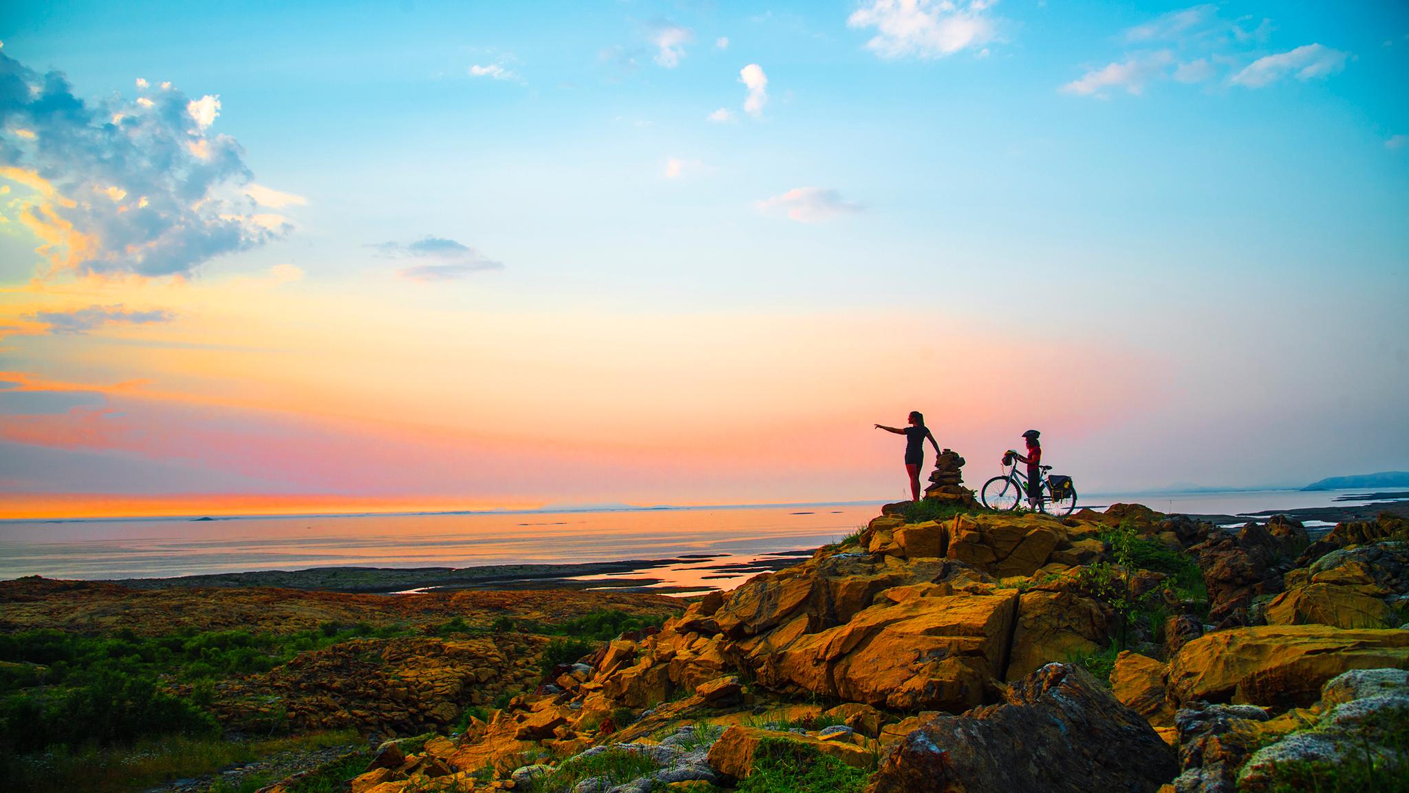 A hiker and a cyclist standing side by side watching the sunset at the Leka island in Trøndelag.