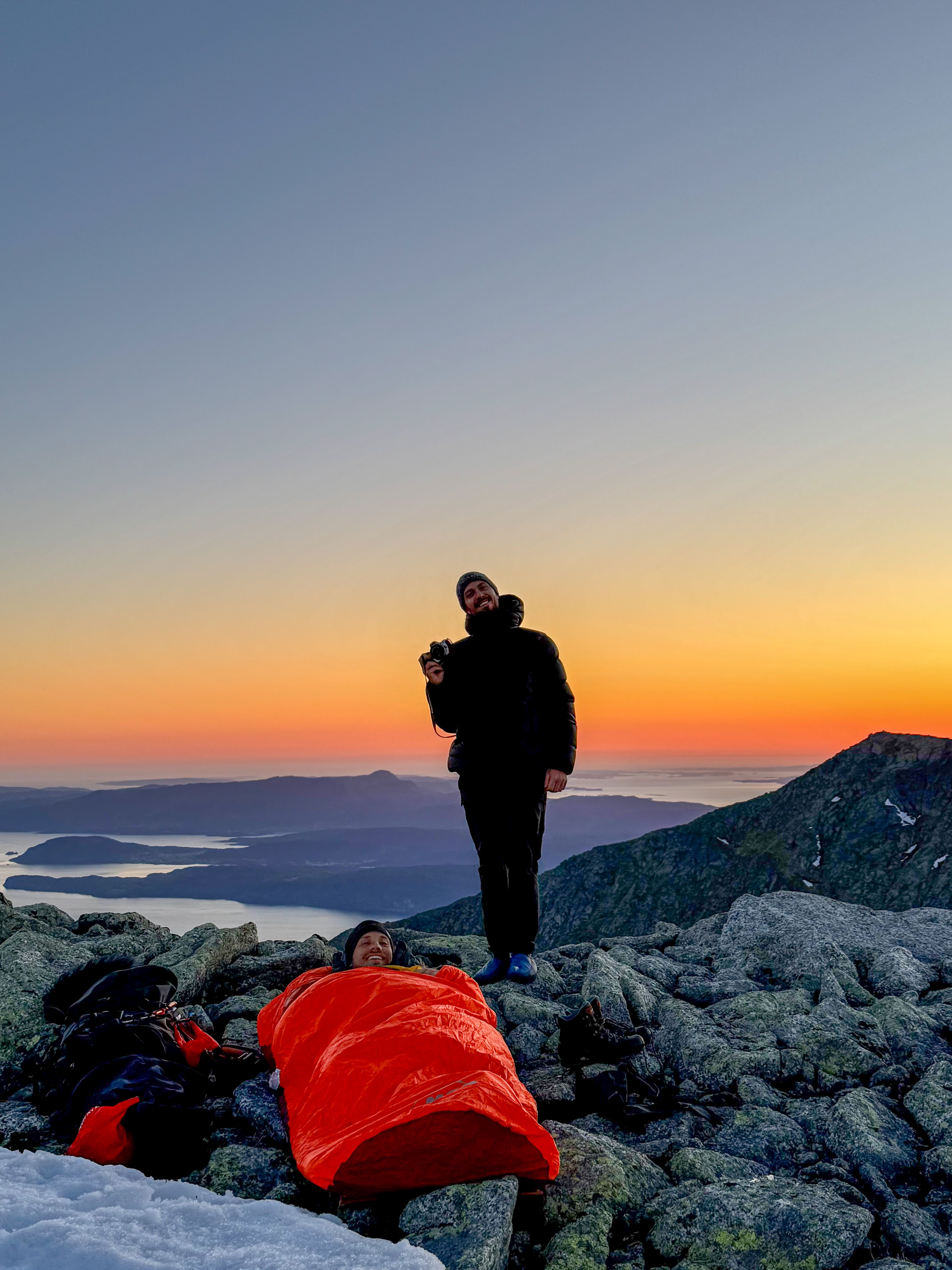 A man standing, and another man is lying in his sleeping bag in the sunset at Indre Laurdalstind in Rosendal in Hardanger