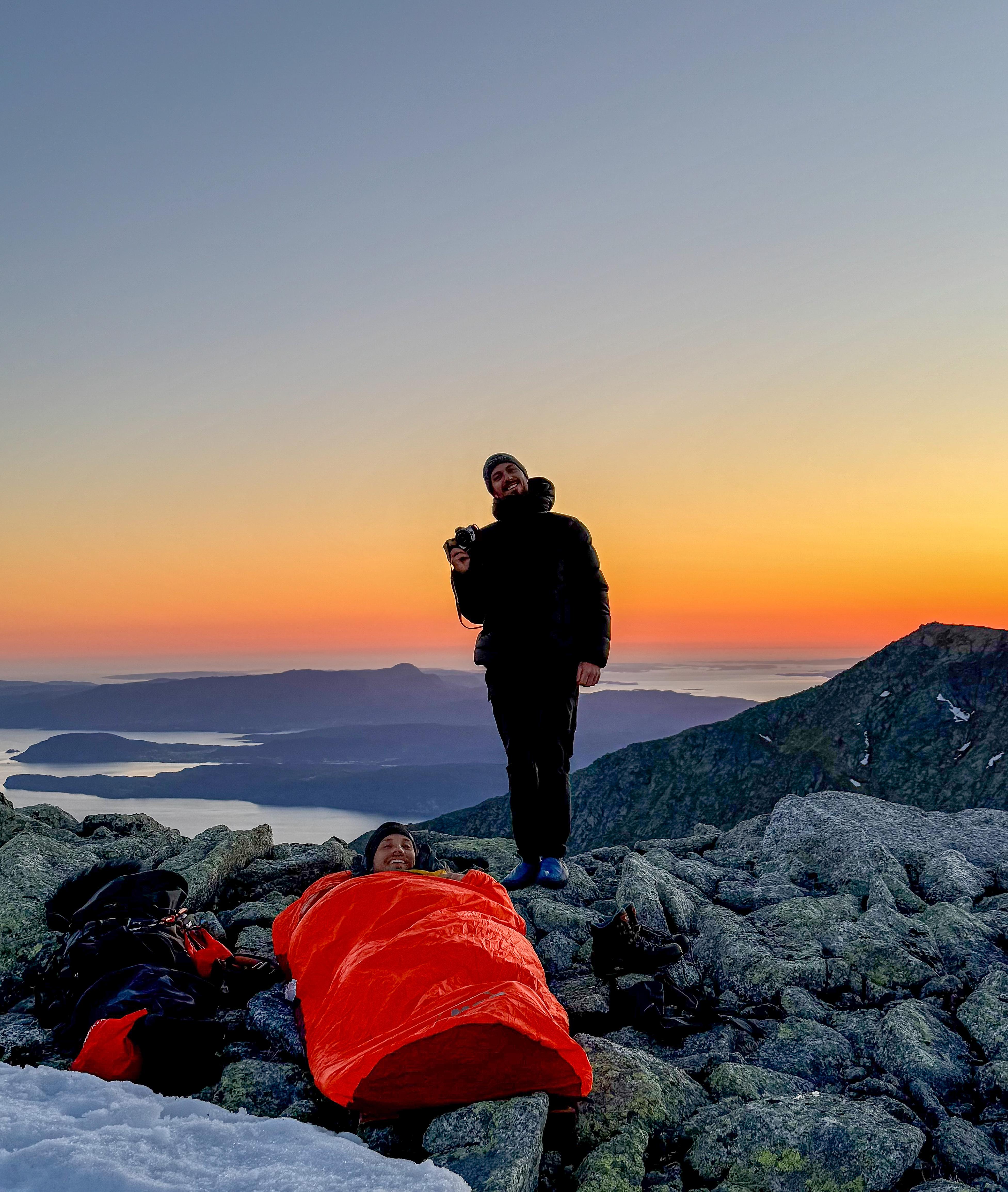 A man standing, and another man is lying in his sleeping bag in the sunset at Indre Laurdalstind in Rosendal in Hardanger