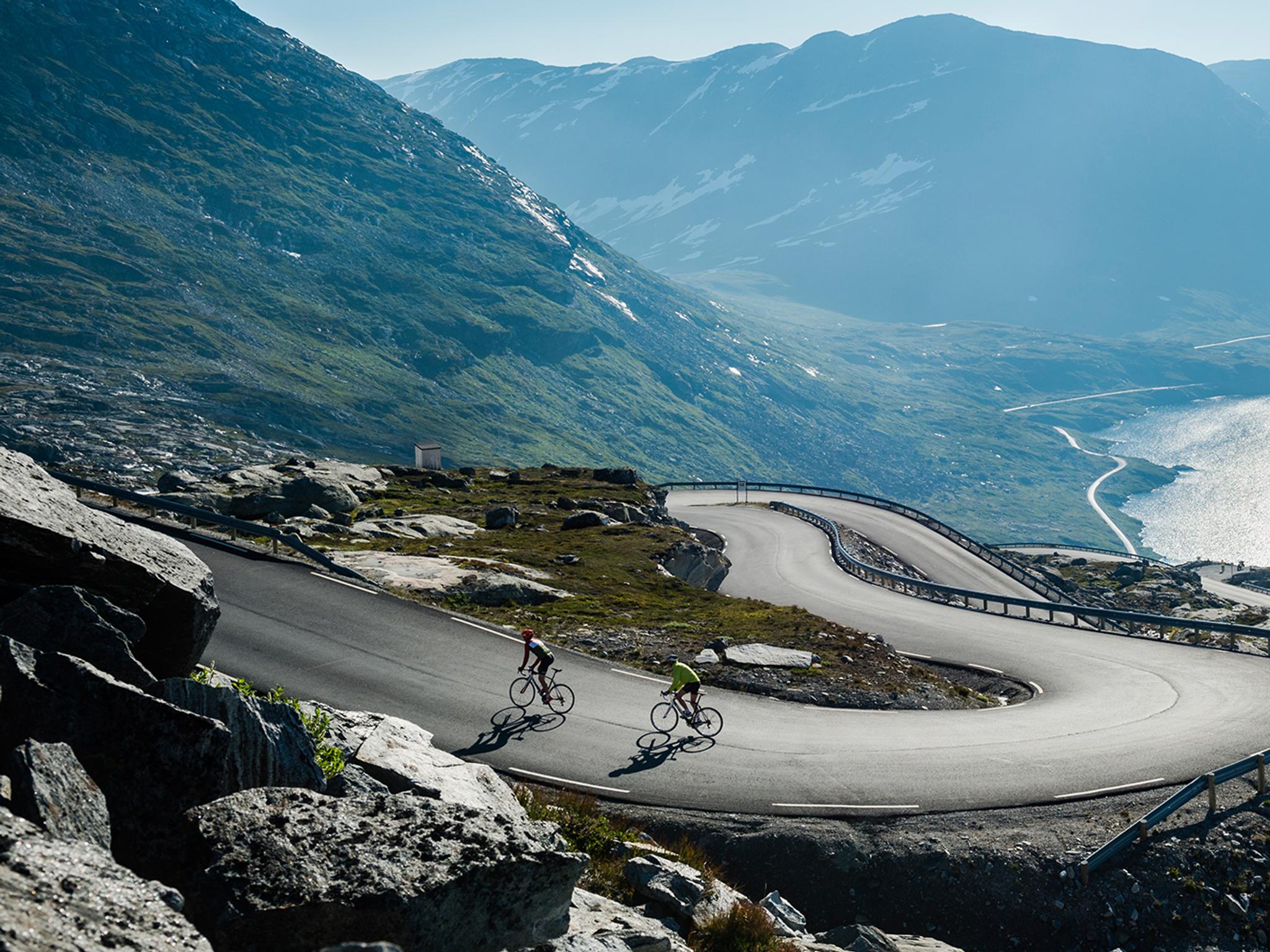 Två personer cyklar längs en slingrande väg ovanför Geirangerfjorden i regionen Fjord Norge (Vestlandet)