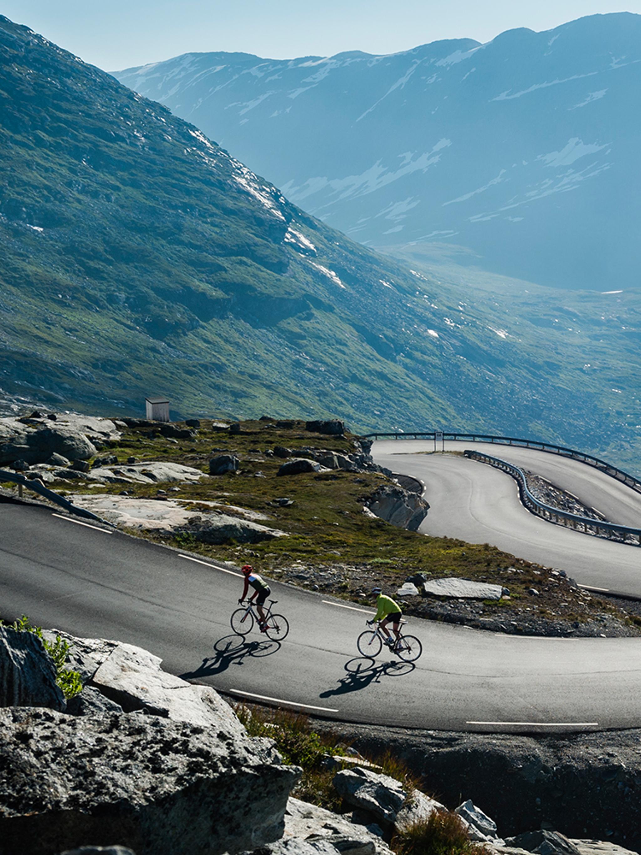 Två personer cyklar längs en slingrande väg ovanför Geirangerfjorden i regionen Fjord Norge (Vestlandet)