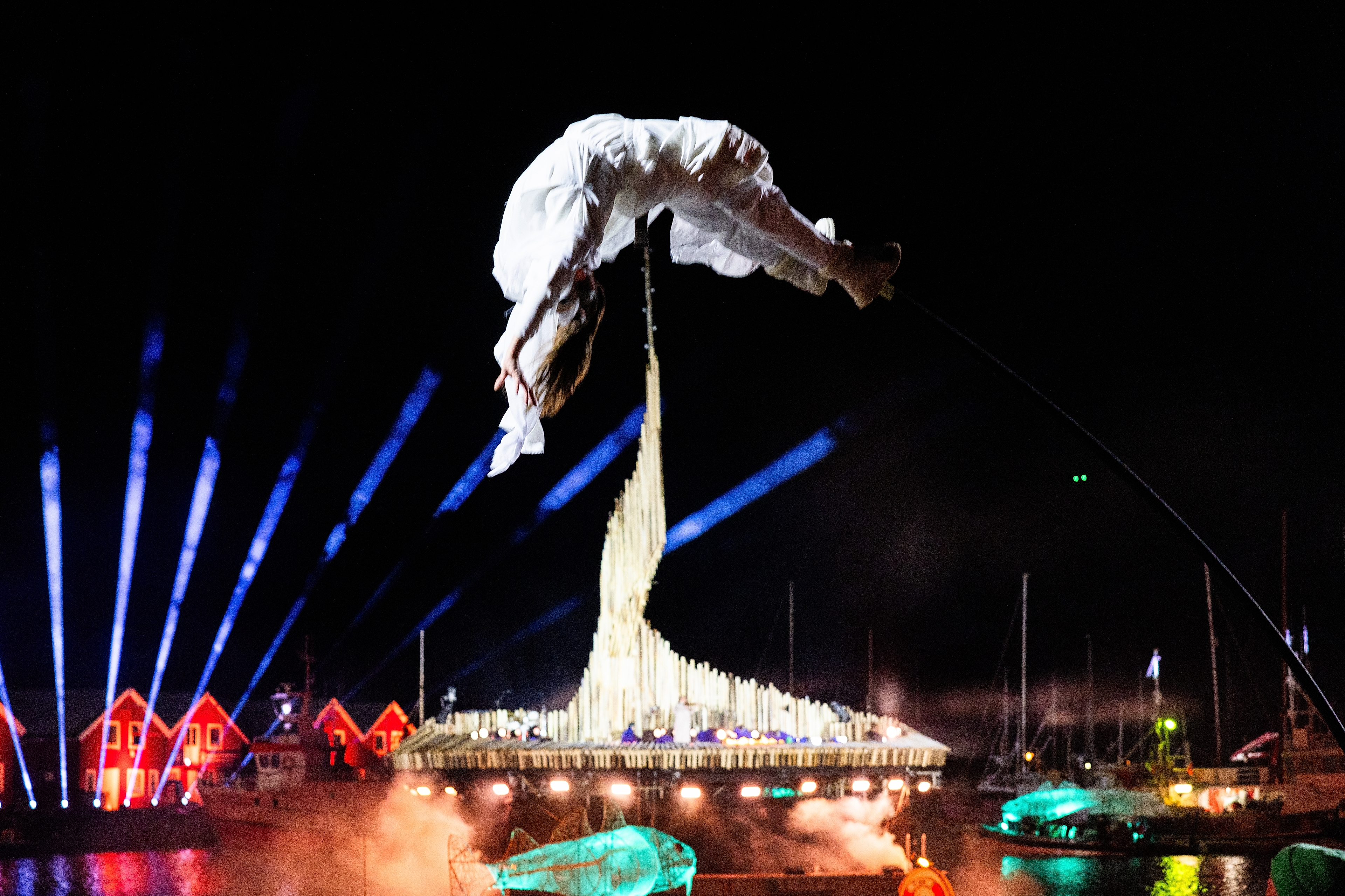 Woman flying on stilts with a lit up stage floating on the sea in the background
