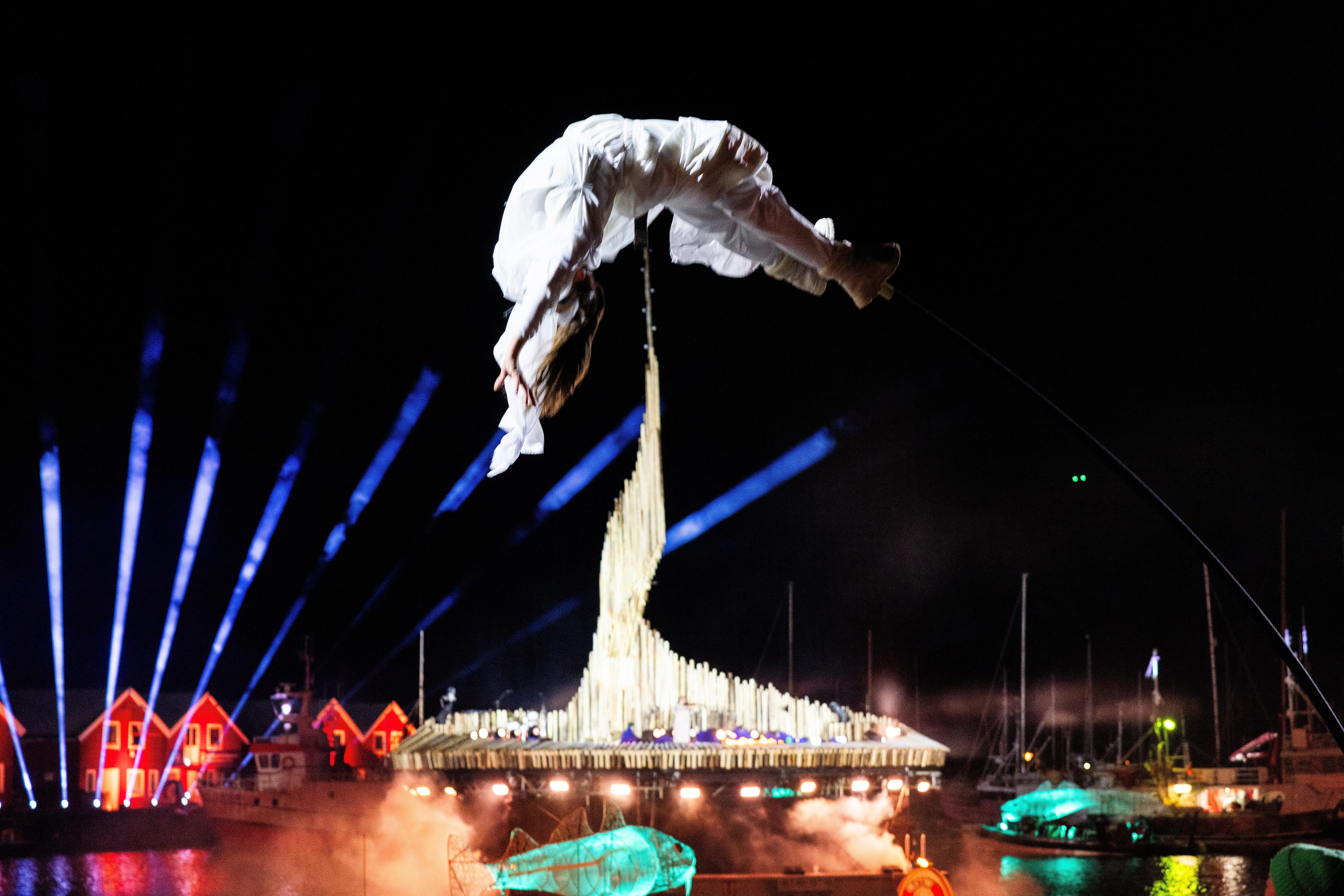 Woman flying on stilts with a lit up stage floating on the sea in the background