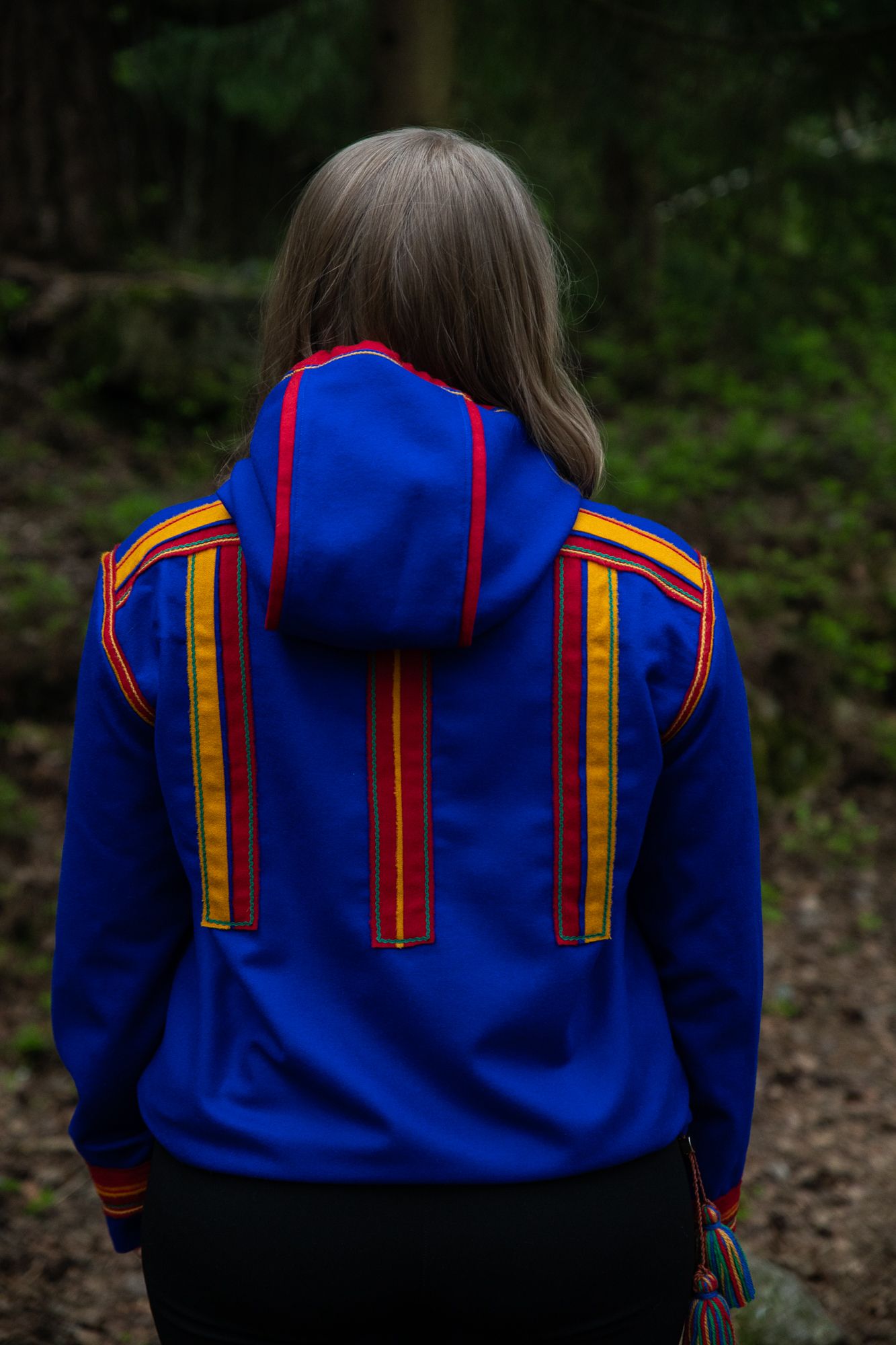 A woman in the woods in a Sami inspired hoodie with her back towards the camera