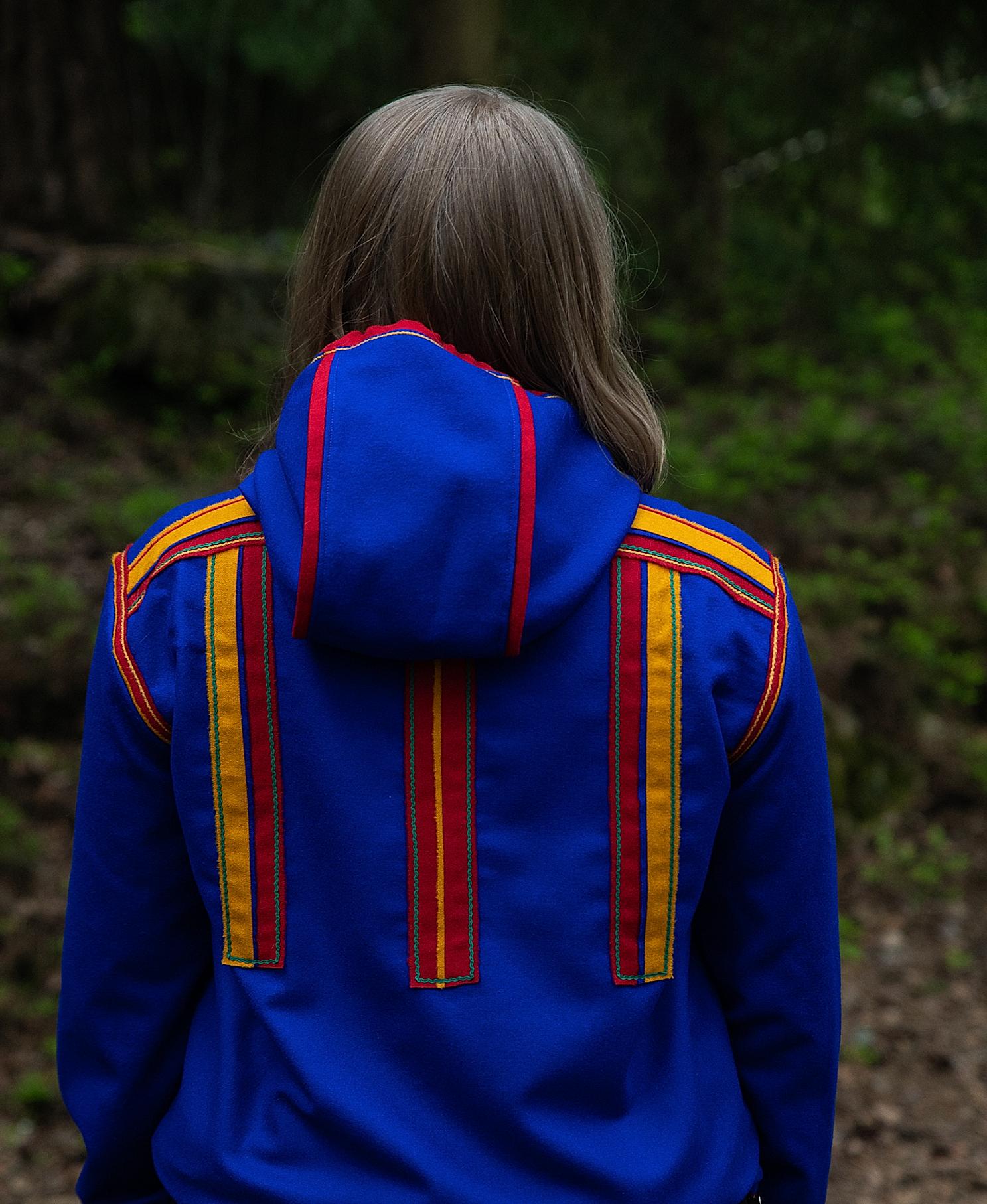 A woman in the woods in a Sami inspired hoodie with her back towards the camera