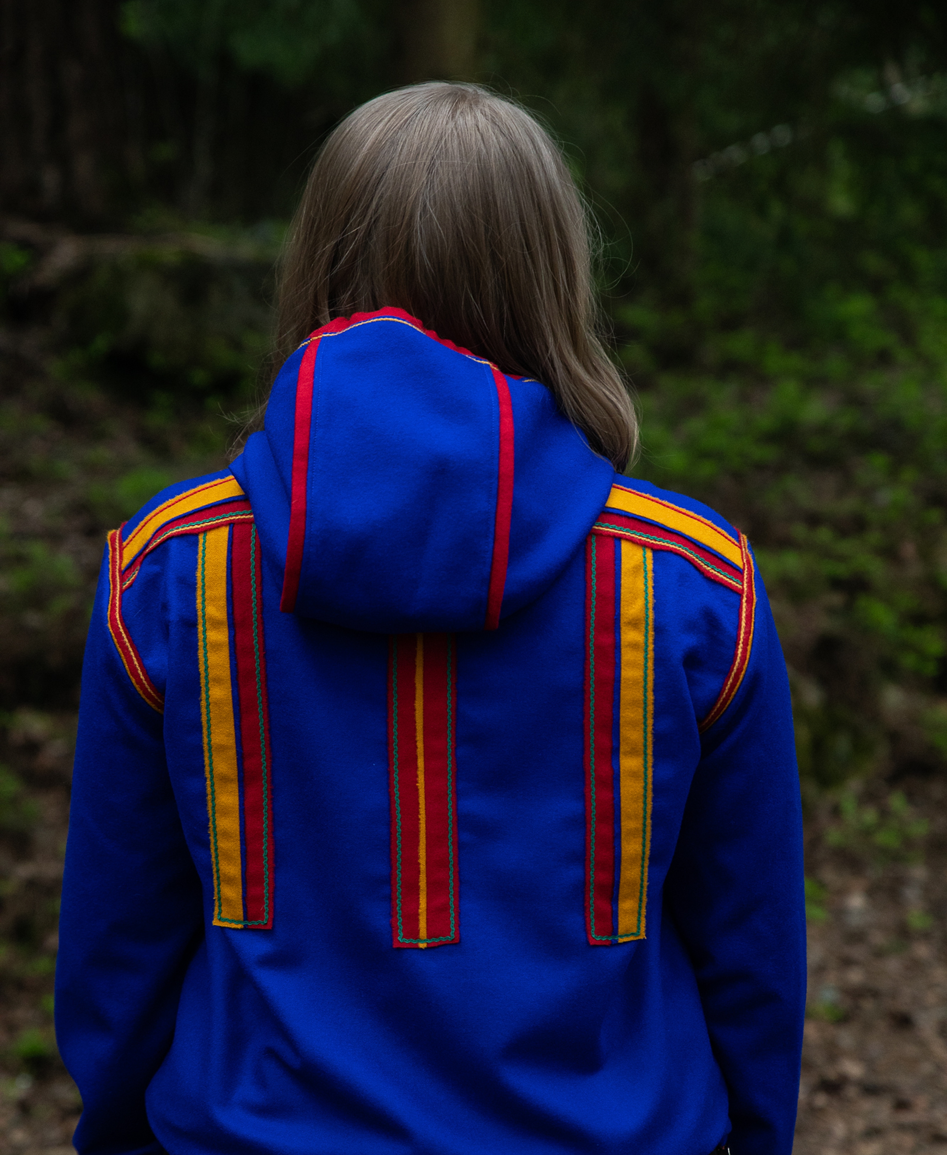 A woman in the woods in a Sami inspired hoodie with her back towards the camera