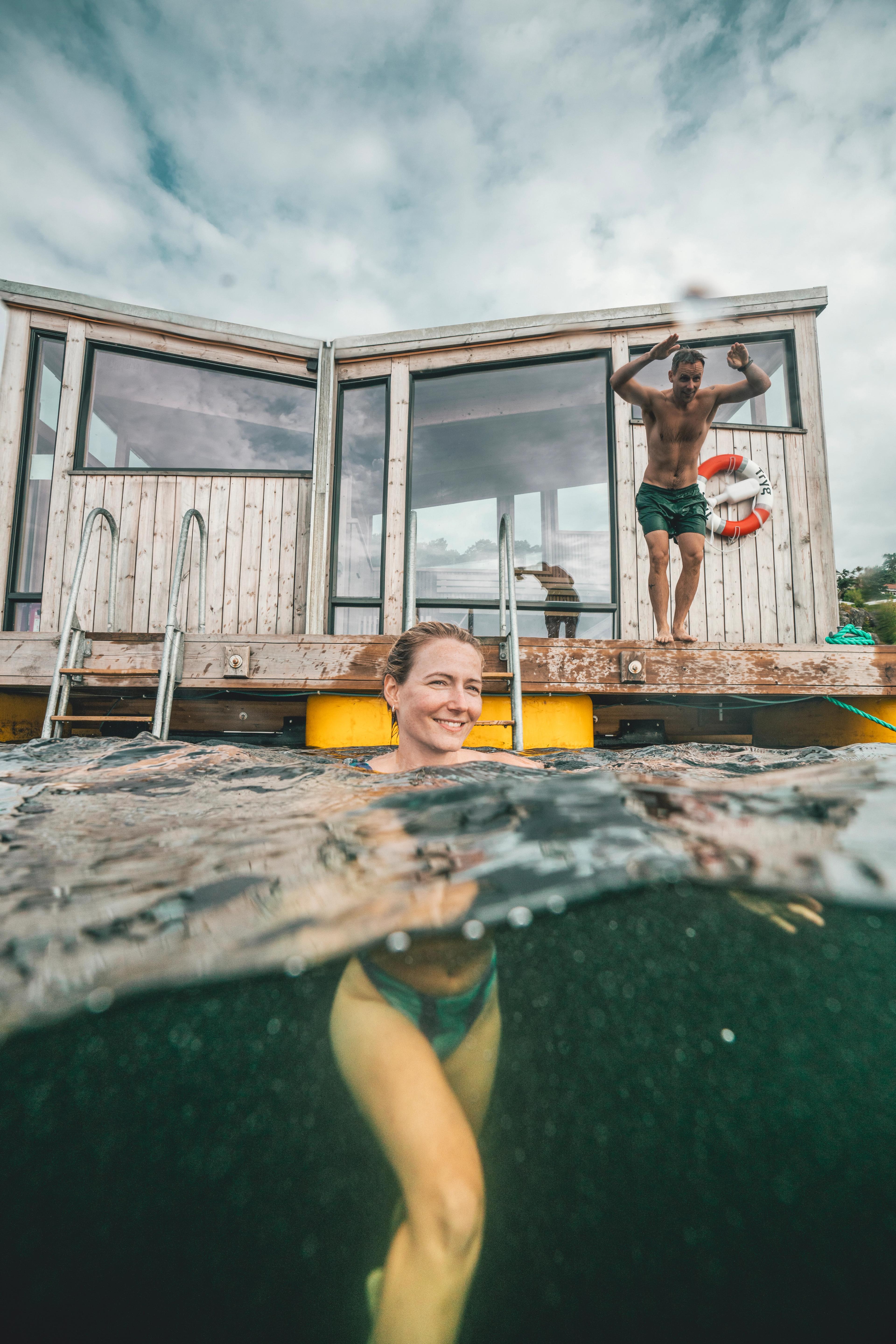 Woman bathing and a man is about to jump in the water in front of the Risør Havbad sauna in Southern Norway