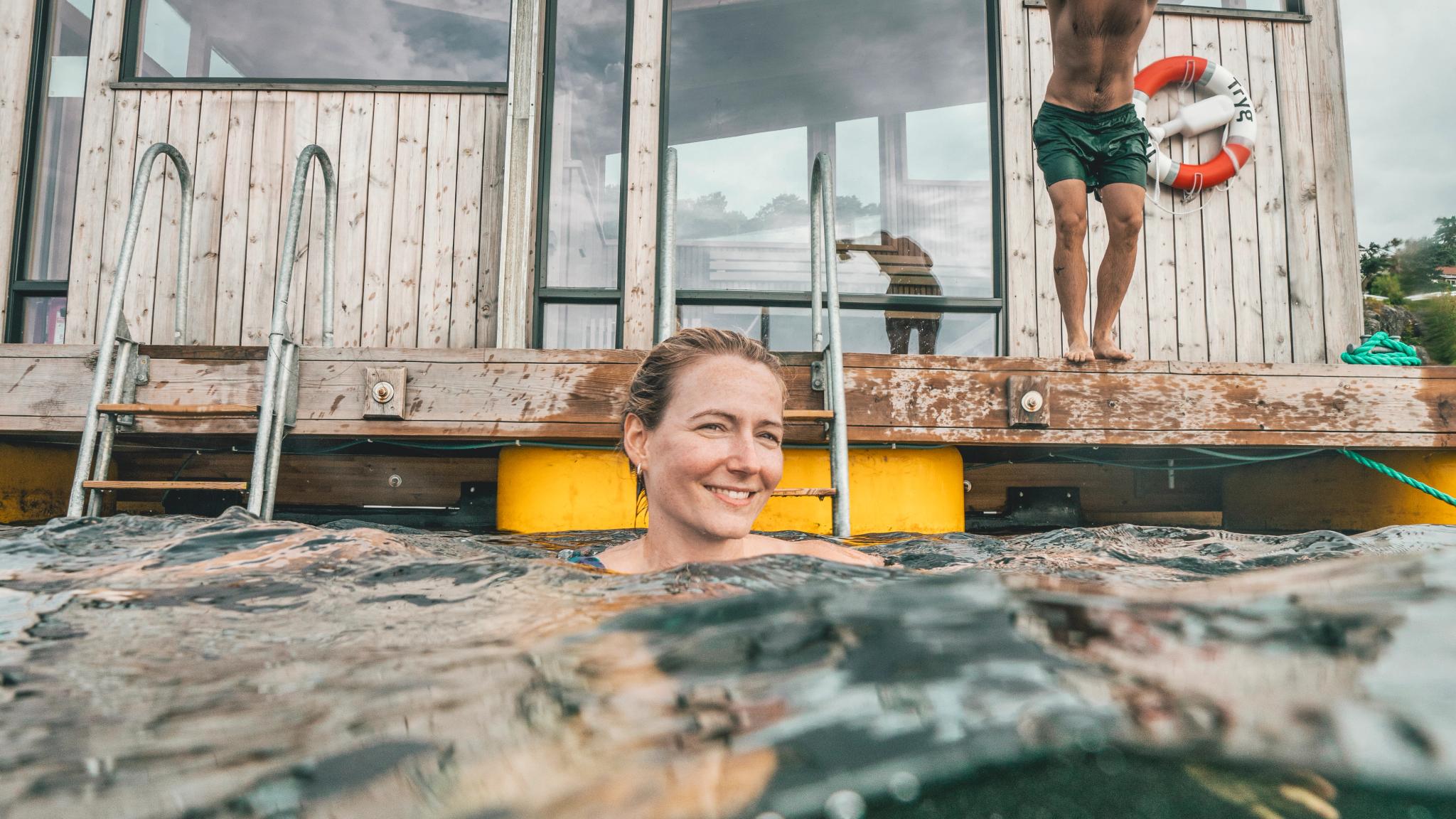 Woman bathing and a man is about to jump in the water in front of the Risør Havbad sauna in Southern Norway