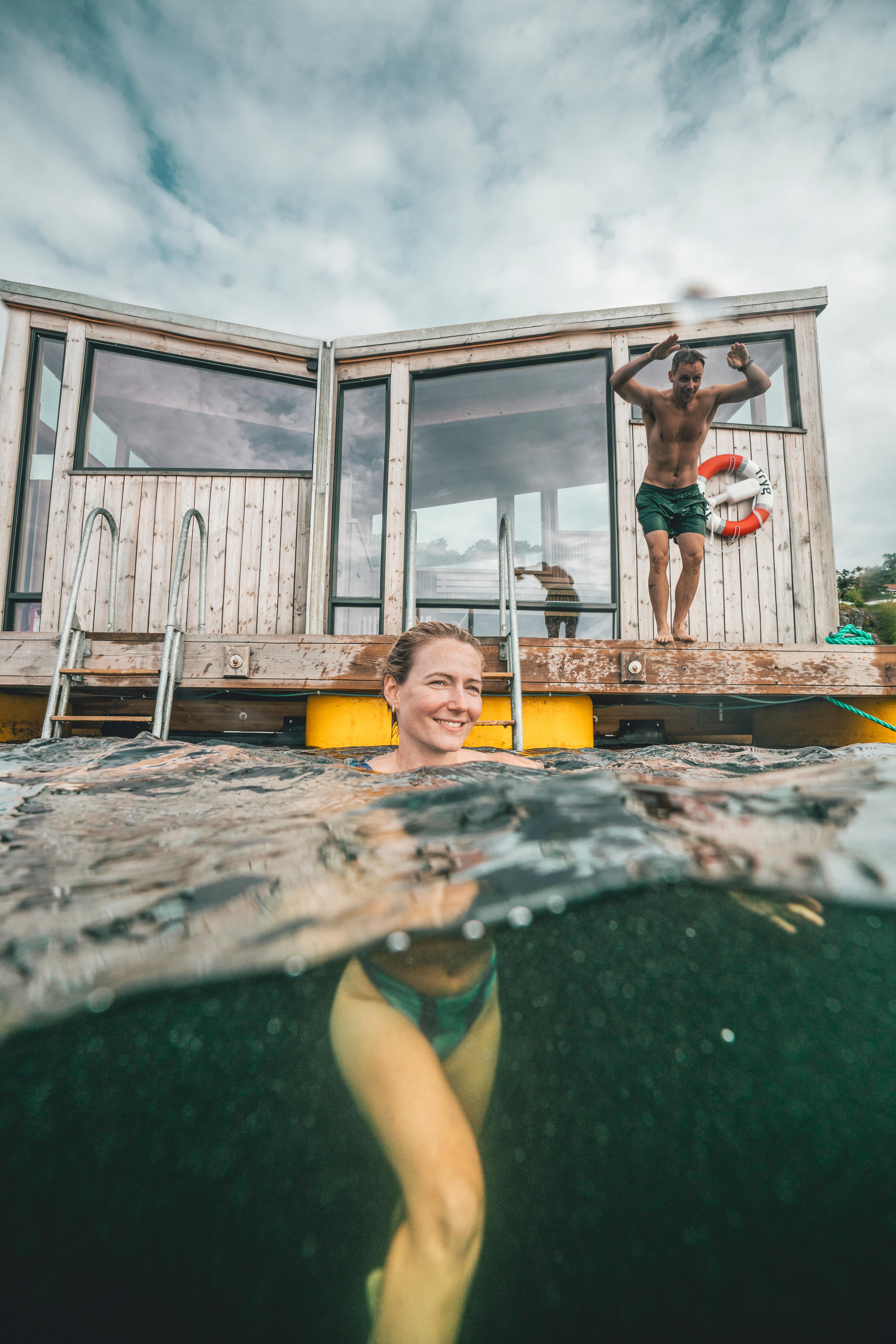 Woman bathing and a man is about to jump in the water in front of the Risør Havbad sauna in Southern Norway