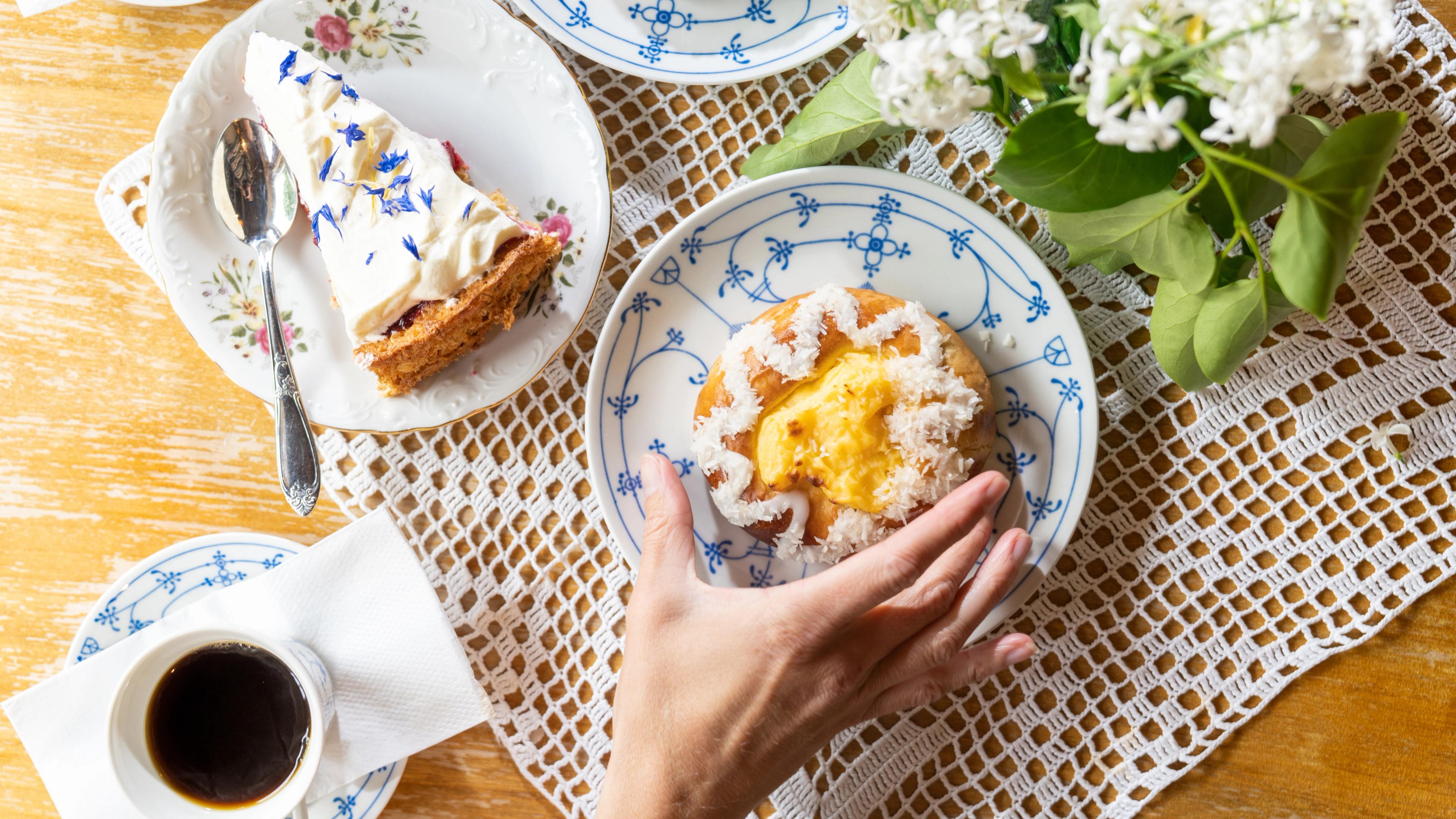 A slice of cake on a plate, a cup of coffee and a skolebrød on a plate with a hand next to it.