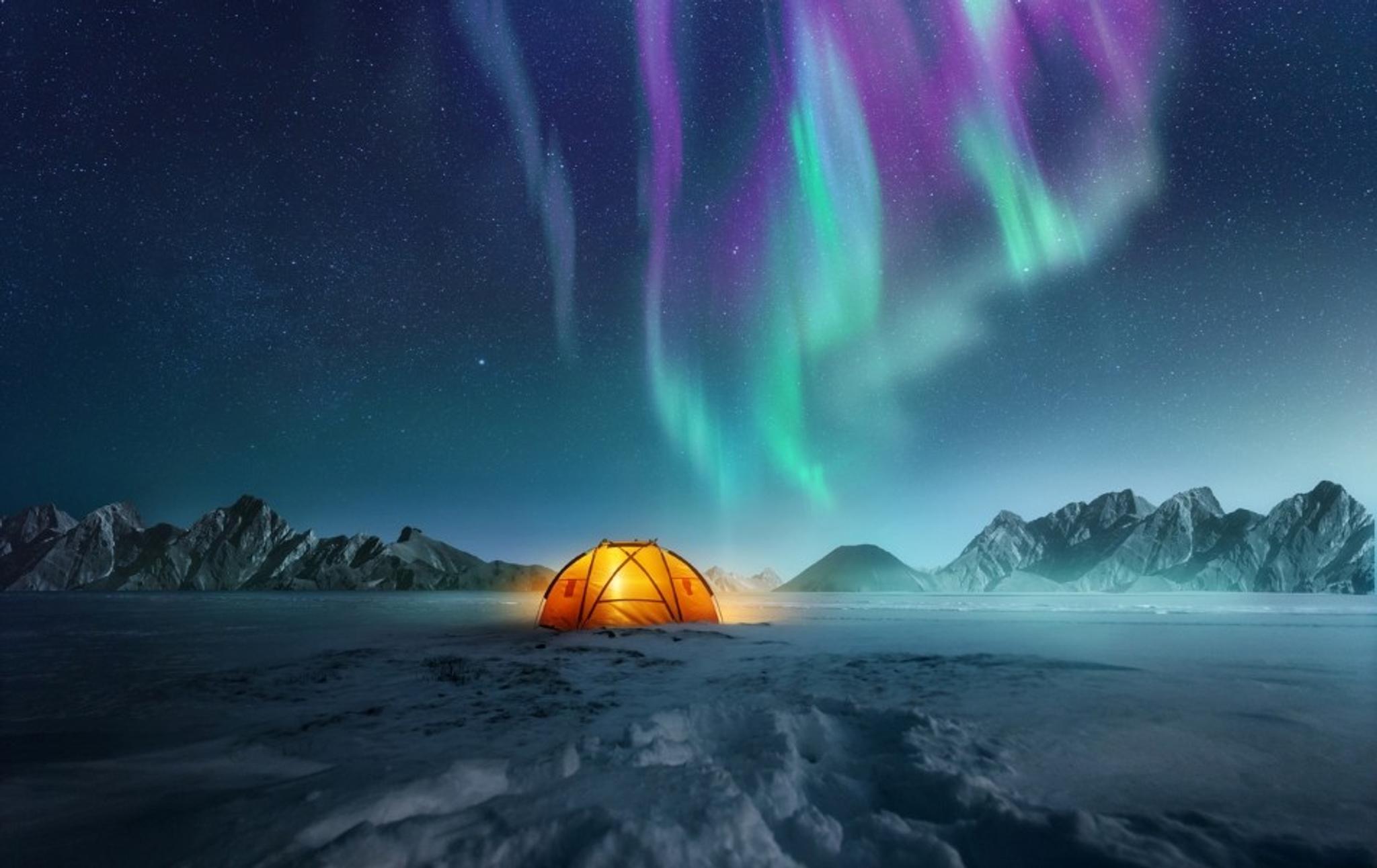 Lit-up tent in snow with colorful northern lights and mountains behind it