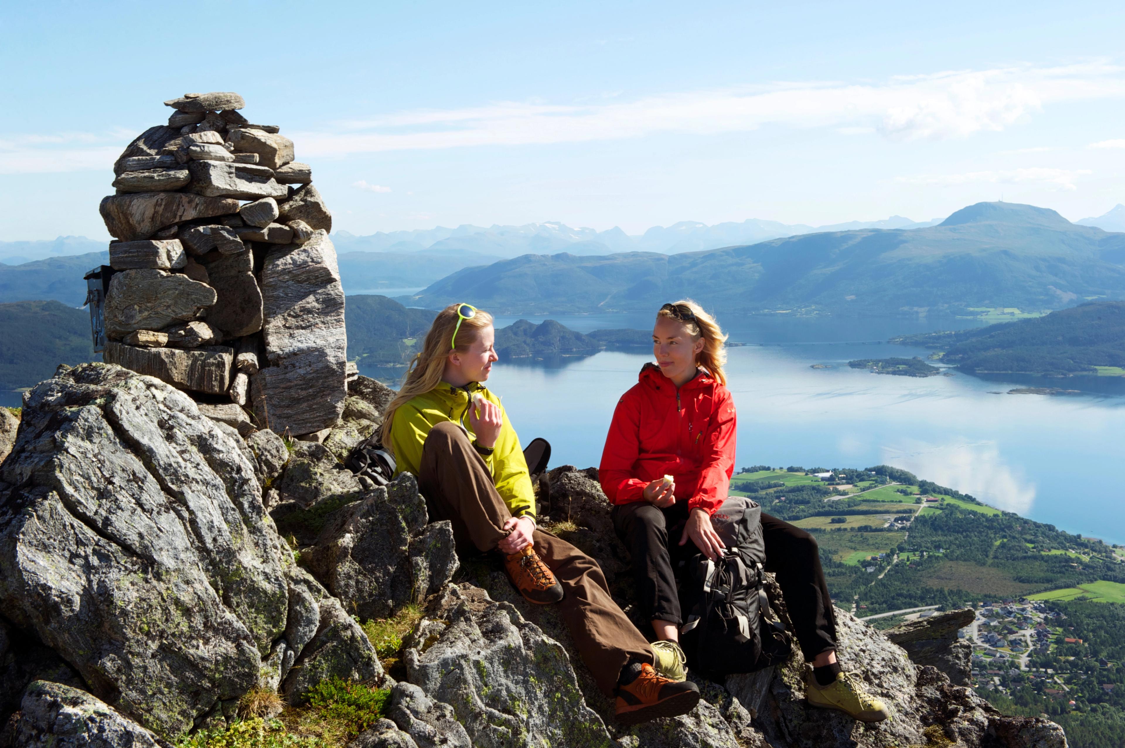 Two women sitting in the sunshine at the top of Freikollen in Kristiansund, with mountains and the fjord in the background. Fjord Norway.