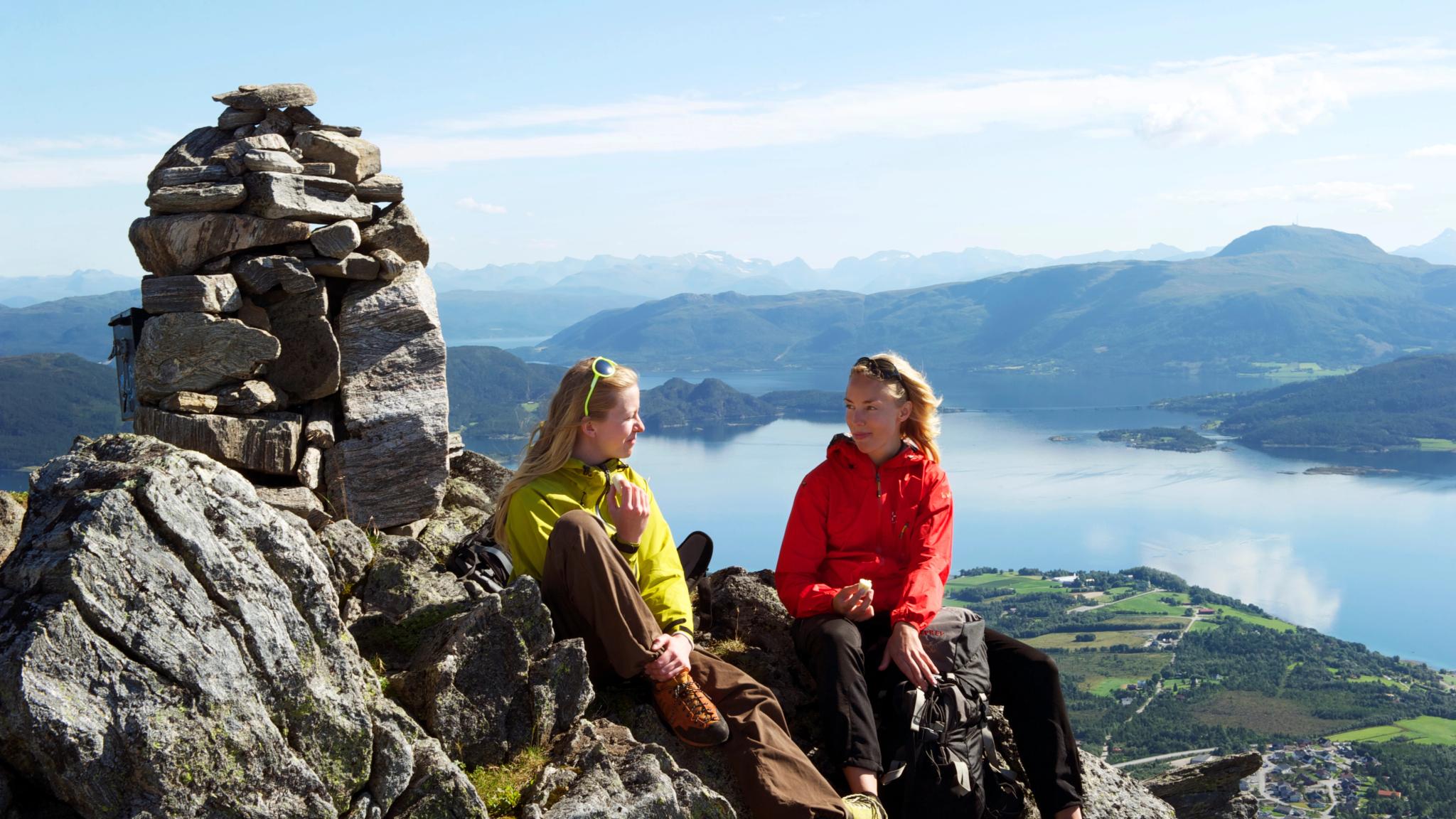 Two women sitting in the sunshine at the top of Freikollen in Kristiansund, with mountains and the fjord in the background. Fjord Norway.