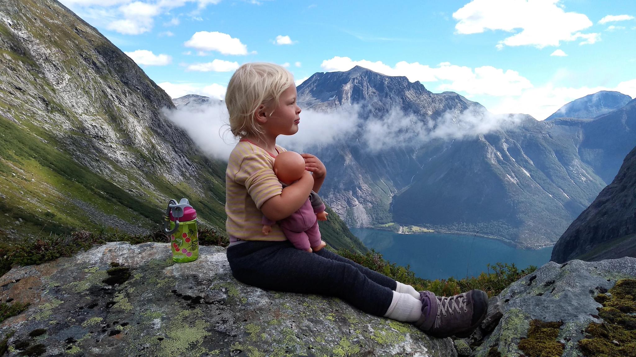 A young girl sitting with her doll surrounded by mountains at Hoemskaret in Romsdalen, Fjord Norway