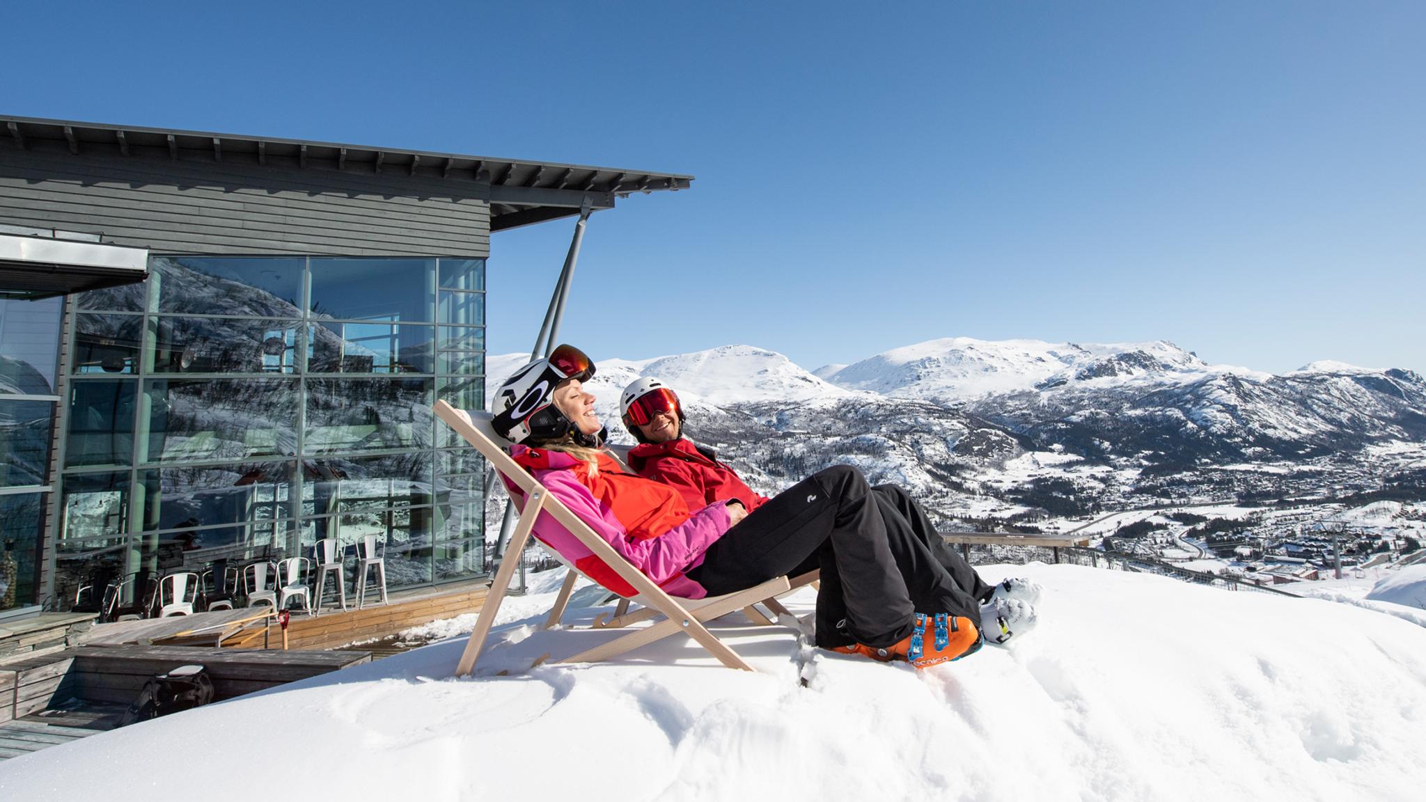 Two people in skiing clothes sit in deck chairs outside Skarsnuten Hotel and enjoy the sunny weather and the wide views of the mountains of Hemsedal, Eastern Norway
