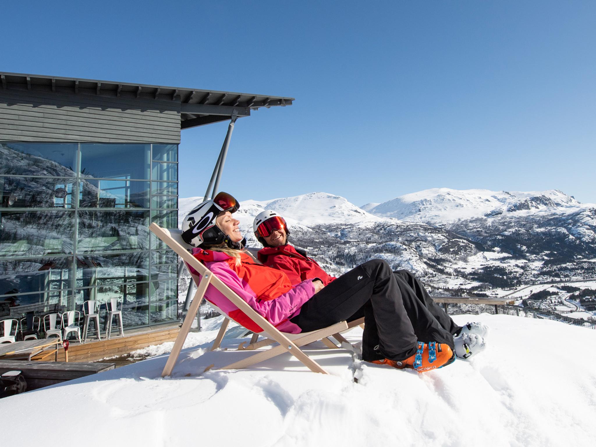 Two people in skiing clothes sit in deck chairs outside Skarsnuten Hotel and enjoy the sunny weather and the wide views of the mountains of Hemsedal, Eastern Norway