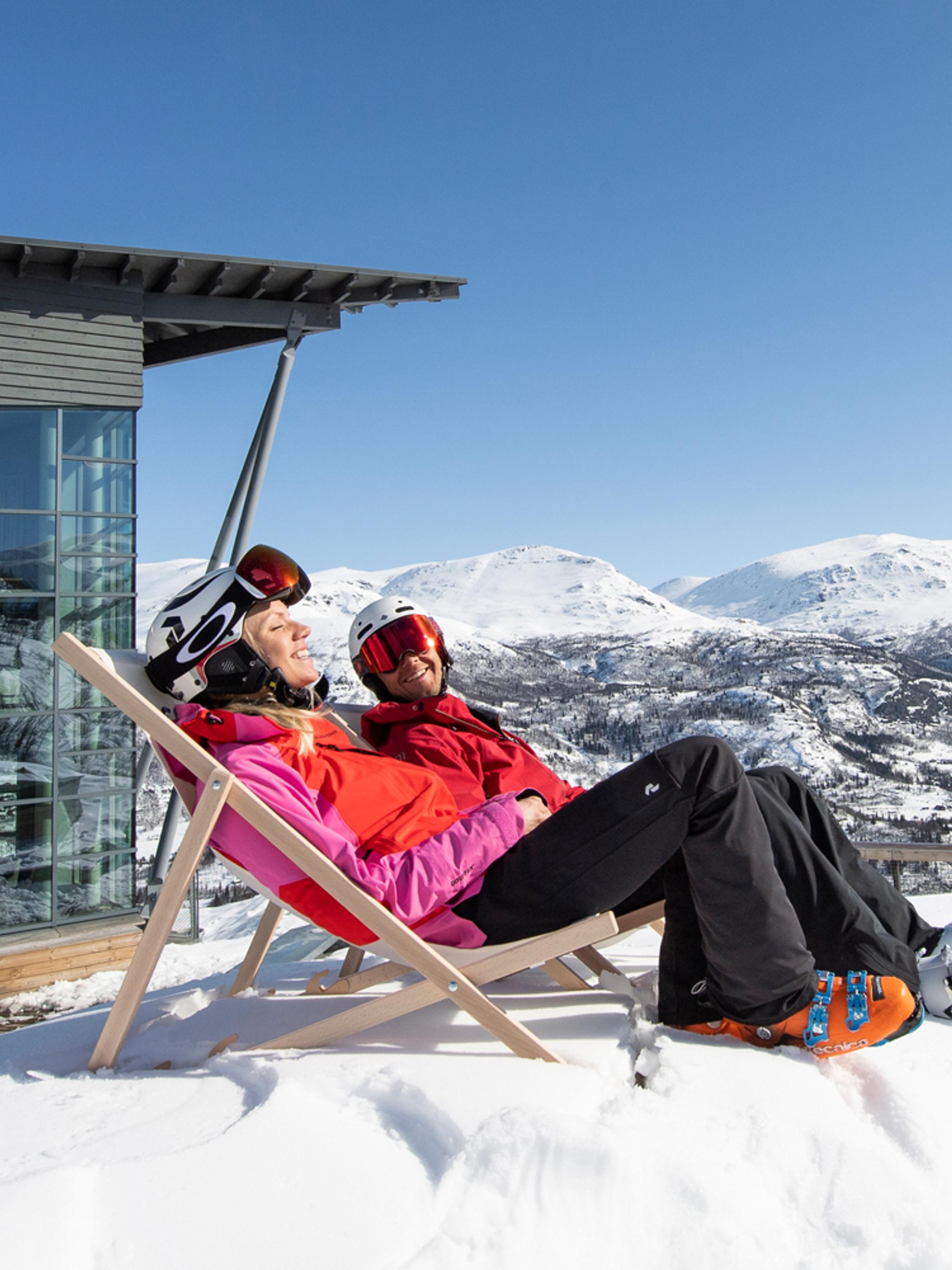Two people in skiing clothes sit in deck chairs outside Skarsnuten Hotel and enjoy the sunny weather and the wide views of the mountains of Hemsedal, Eastern Norway