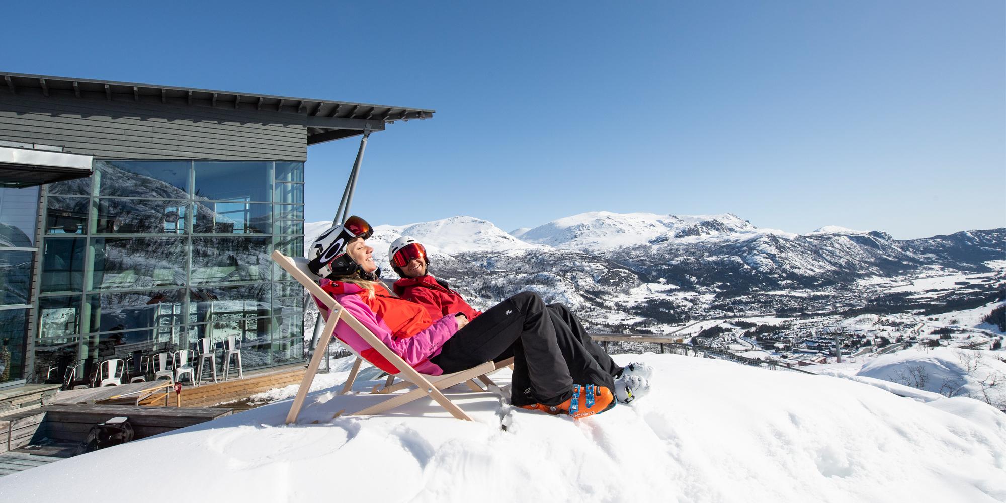 Two people in skiing clothes sit in deck chairs outside Skarsnuten Hotel and enjoy the sunny weather and the wide views of the mountains of Hemsedal, Eastern Norway