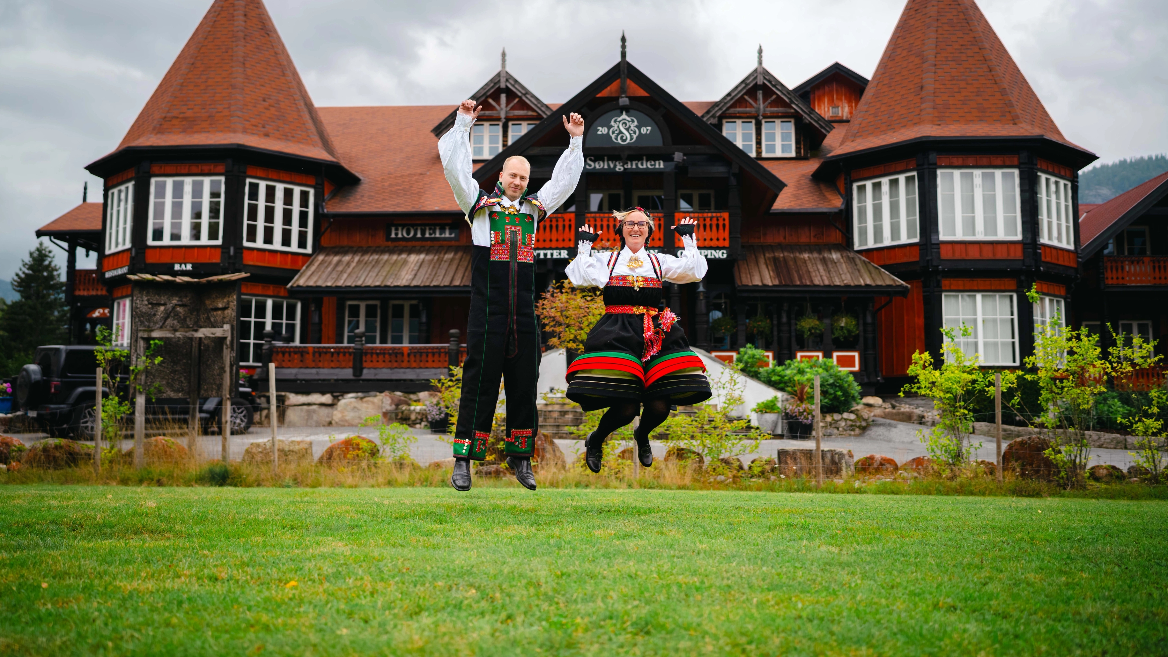 A man and woman jumping joyfully in traditional Setesdal bunads in front of the Sølvgården hotel in the Setesdalen valley