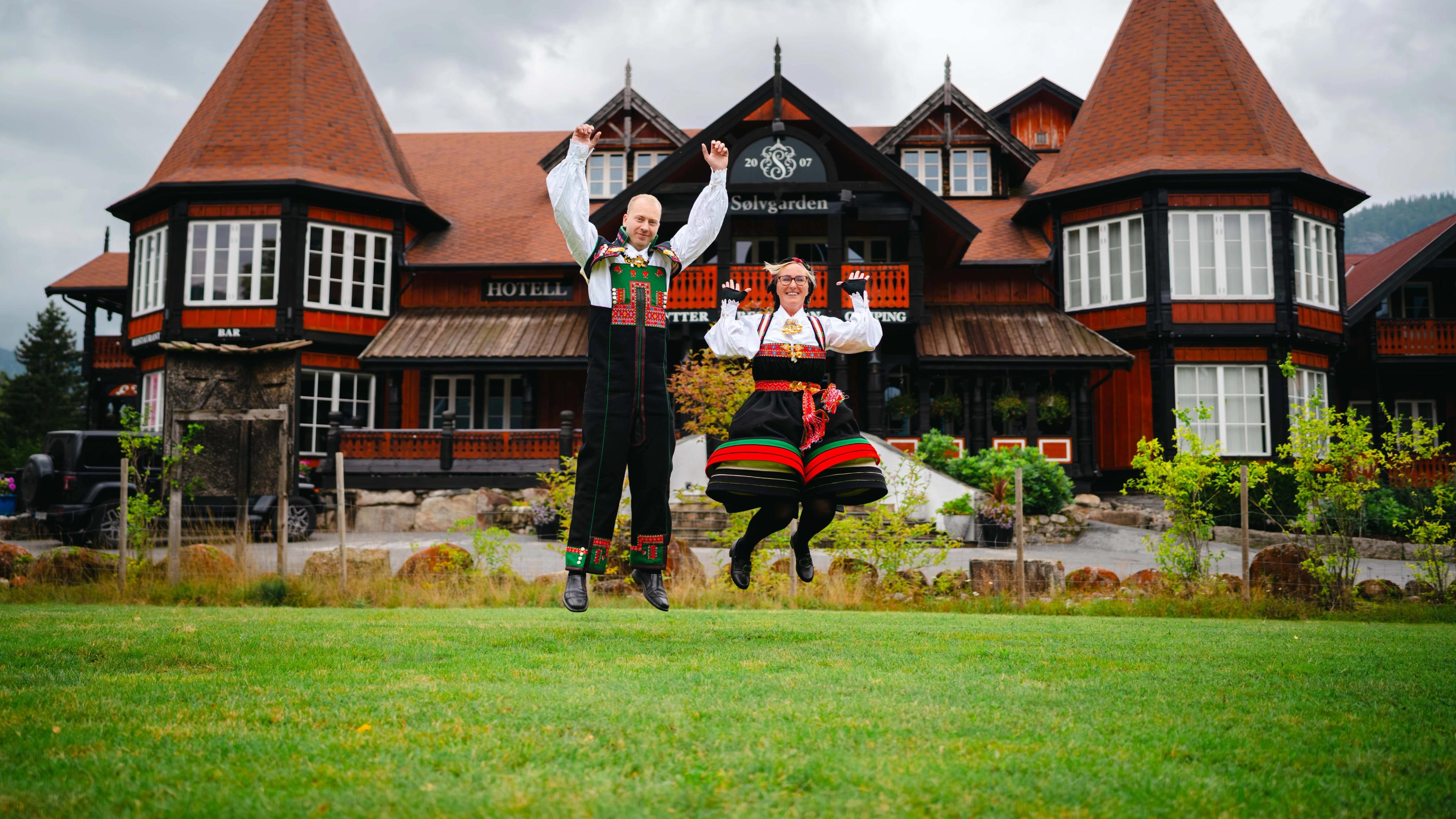 A man and woman jumping joyfully in traditional Setesdal bunads in front of the Sølvgården hotel in the Setesdalen valley