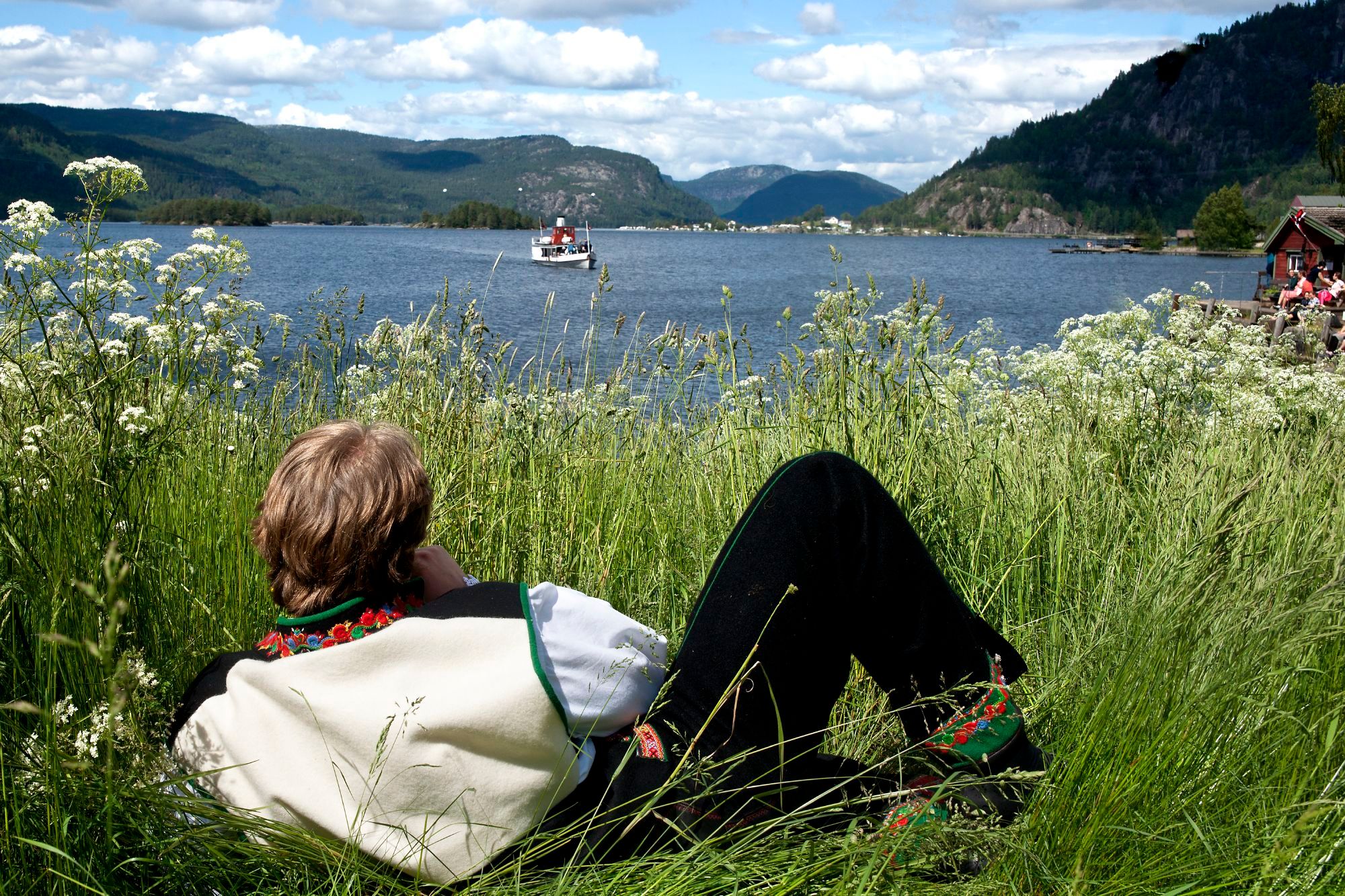 En man klädd i Setesdalsbunad (den lokala nationaldräkten) ligger i gräset. I bakgrunden syns en ångbåt på Byglandsfjorden i Setesdal, södra Norge