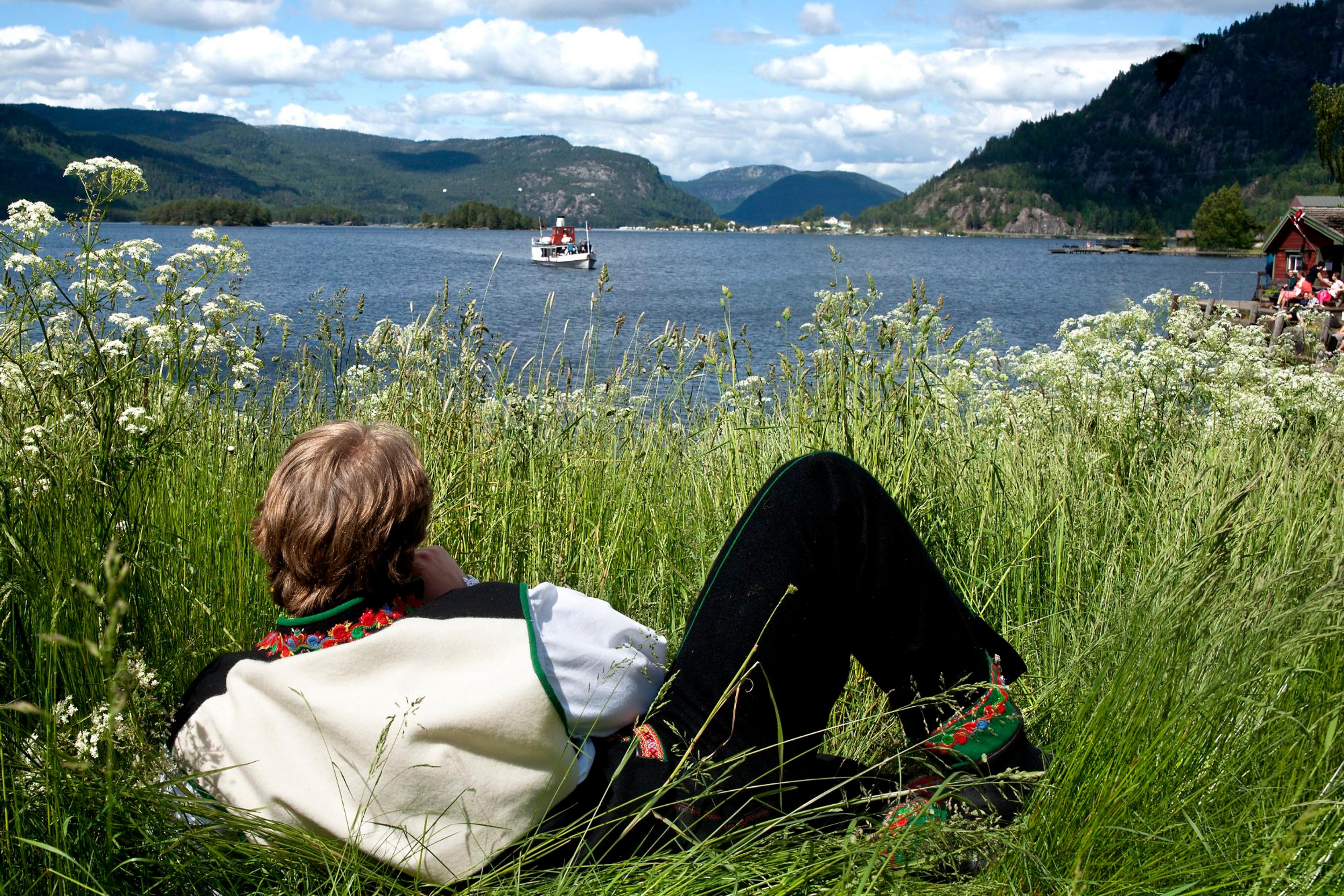 Een man in de nationale klederdracht Setesdalsbunad die in het gras ligt en een oude stoomboot op het Byglandsfjord bekijkt in Setesdal, Zuid-Noorwegen