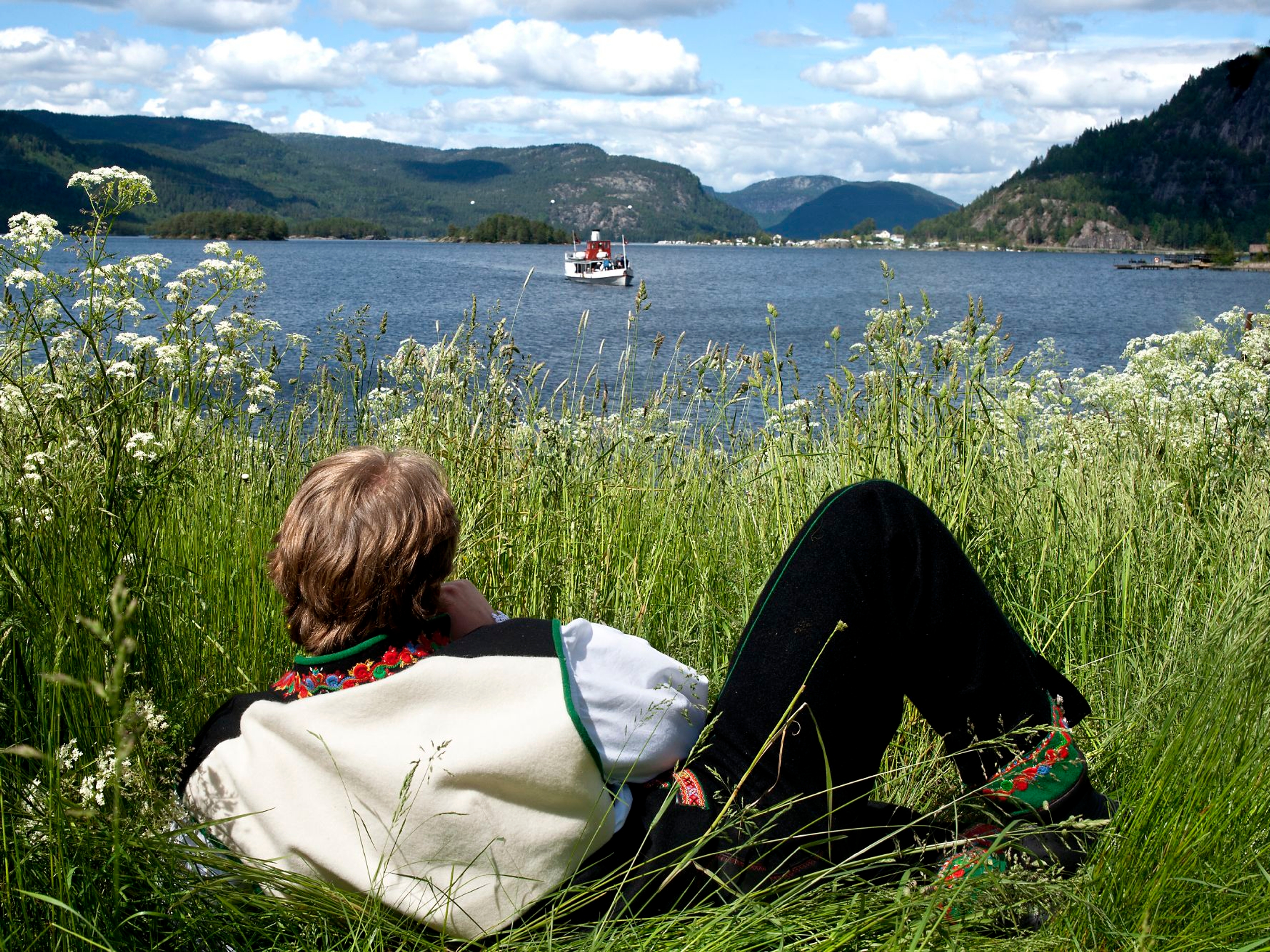 A man in the national costume Setesdalsbunad lying in the grass and watching an old steamboat on the Byglandsfjord in Setesdal, Southern Norway