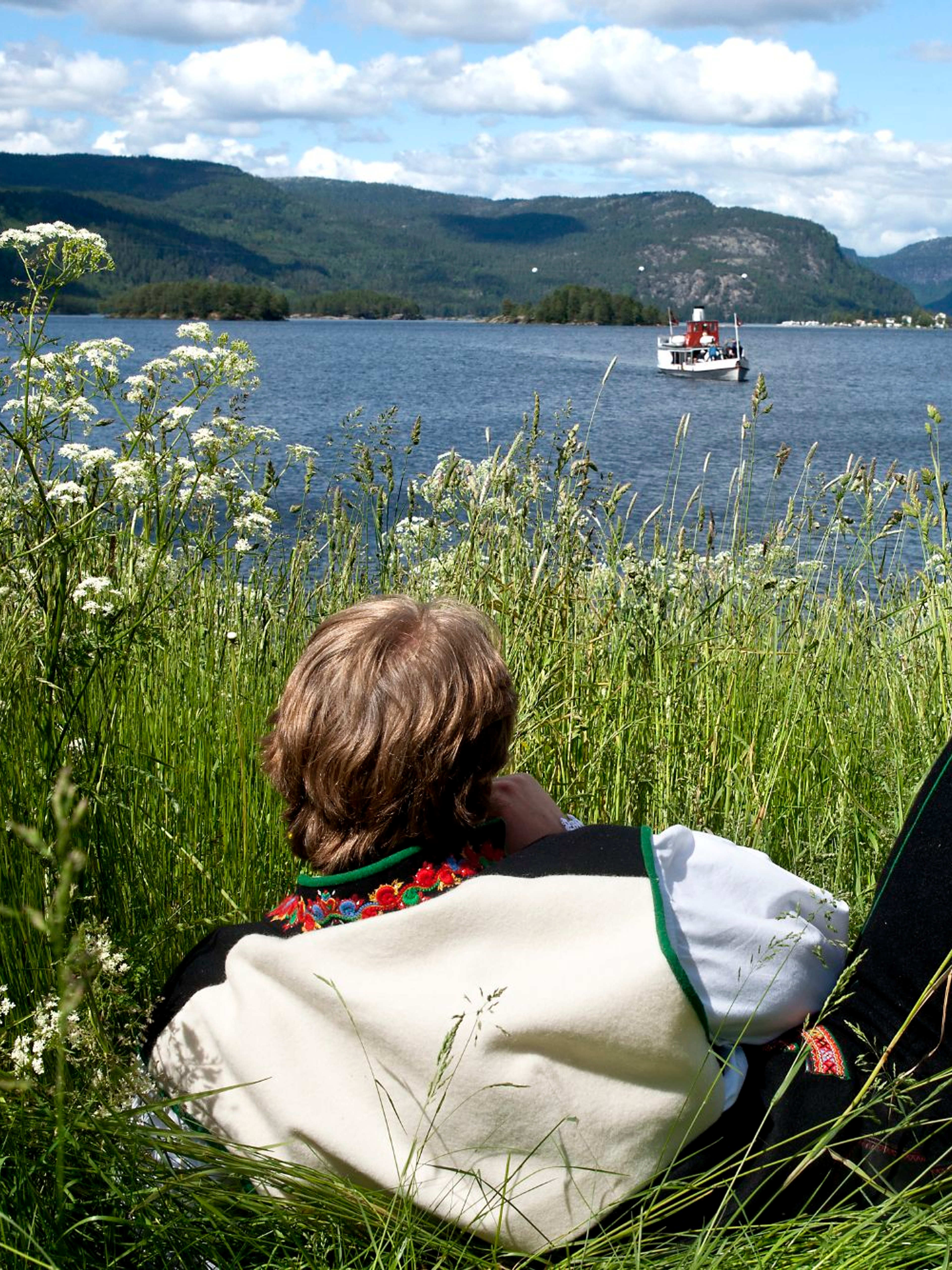 Ein in der Nationaltracht Setesdalbunad liegt im Gras und und schaut auf ein Boot am Byglandsfjord, Setesdal