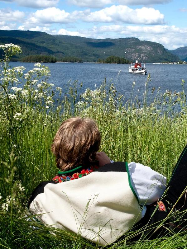 Un hombre ataviado con el típico traje Setesdalsbunad y tumbado sobre la hierba a orillas del Byglandsfjord, en Setesdal, observa cómo un viejo barco de vapor navega sobre el fiordo.