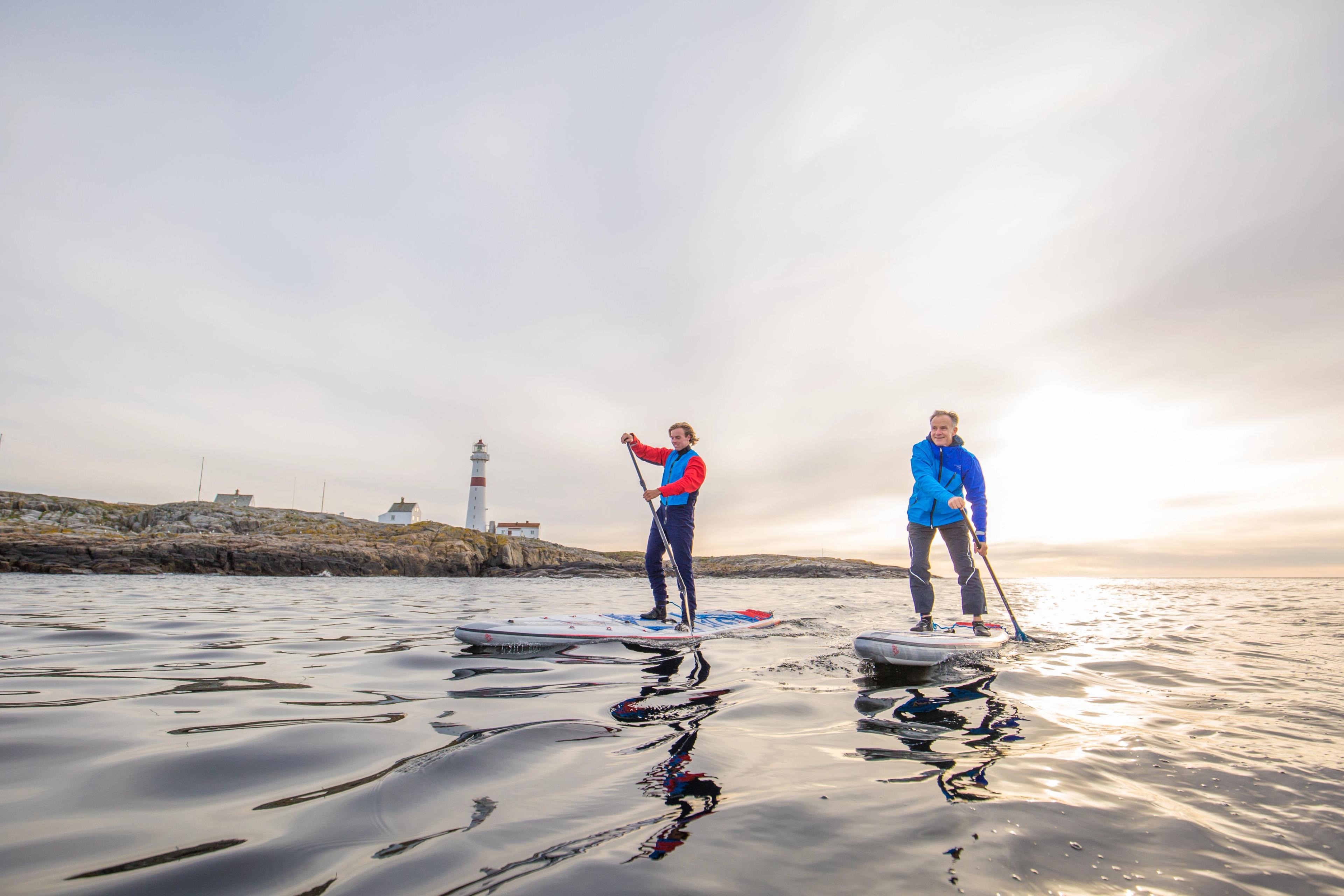 Two people stand up paddleboarding in Arendal, Southern Norway, with Torungen lighthouse in the background.