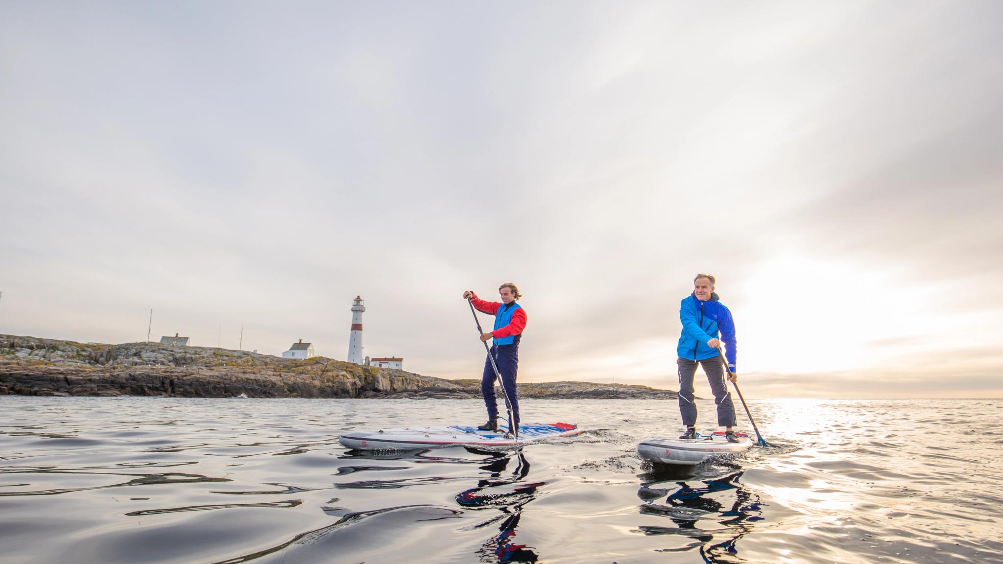 Two people stand up paddleboarding in Arendal, Southern Norway, with Torungen lighthouse in the background.