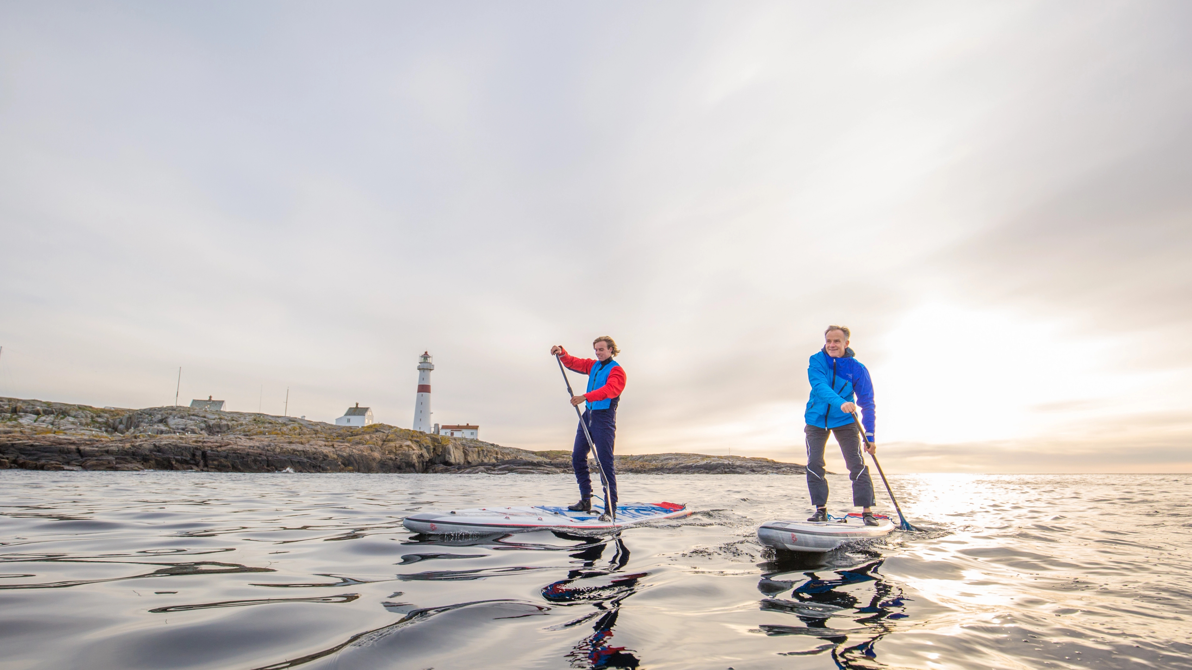 Two people stand up paddleboarding in Arendal, Southern Norway, with Torungen lighthouse in the background.