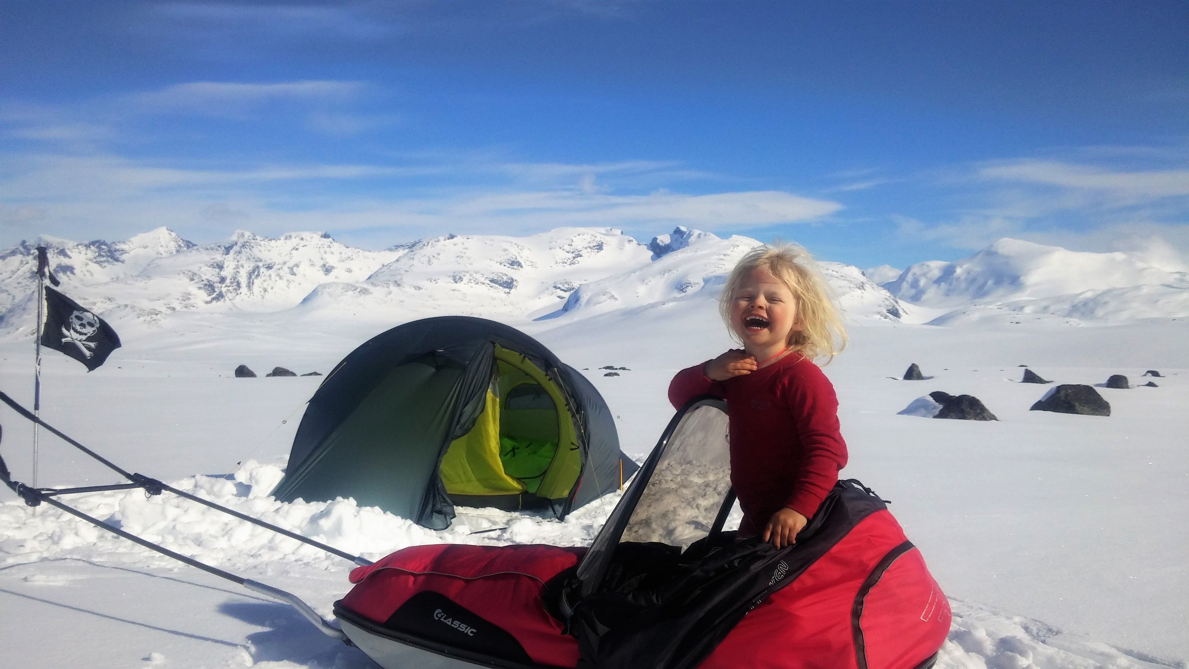 4 år gamla Mina står på en pulka framför ett tält en solig vinterdag i Jotunheimen nationalpark i regionen Østlandet (östra Norge)
