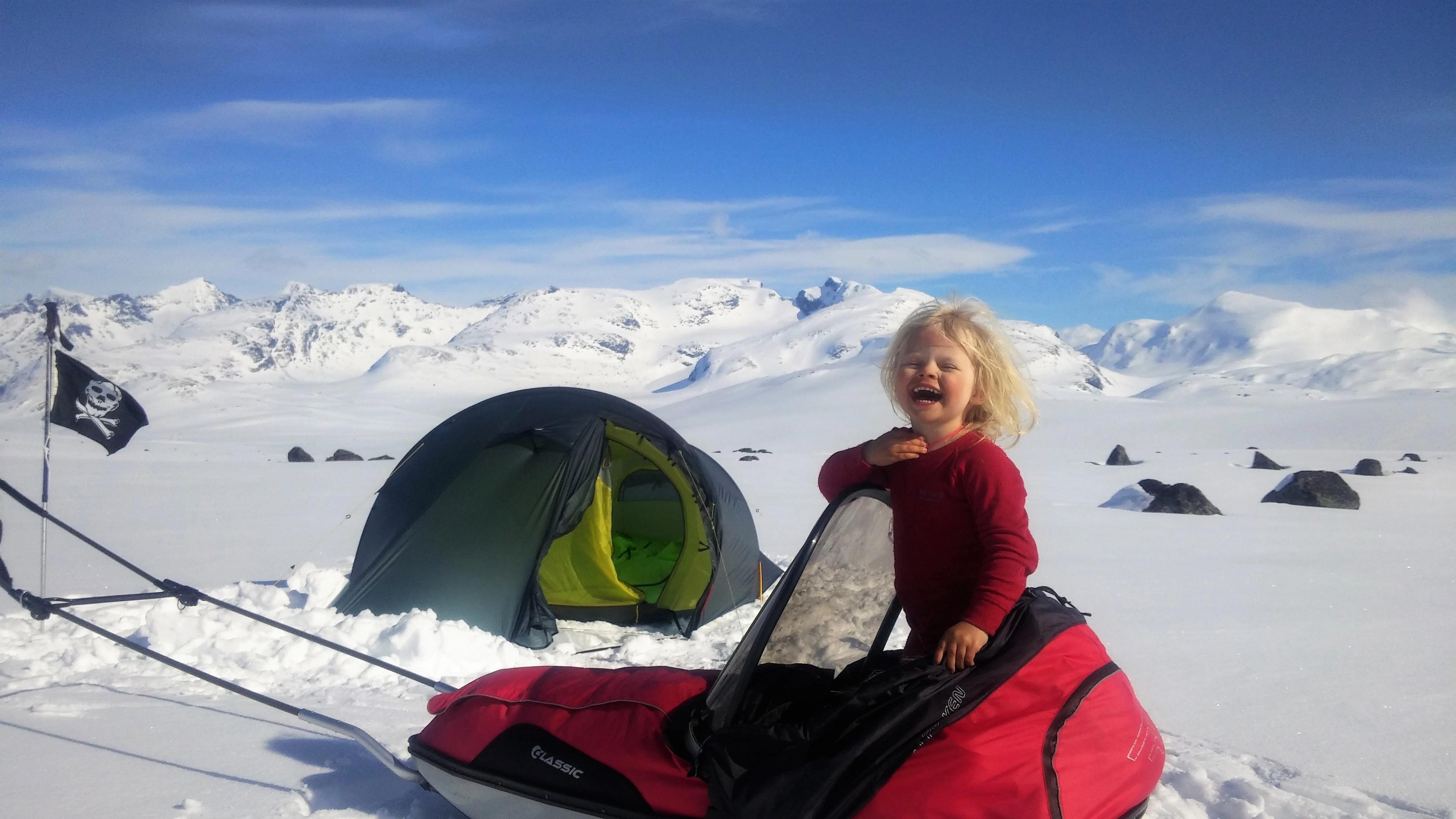 4 år gamla Mina står på en pulka framför ett tält en solig vinterdag i Jotunheimen nationalpark i regionen Østlandet (östra Norge)