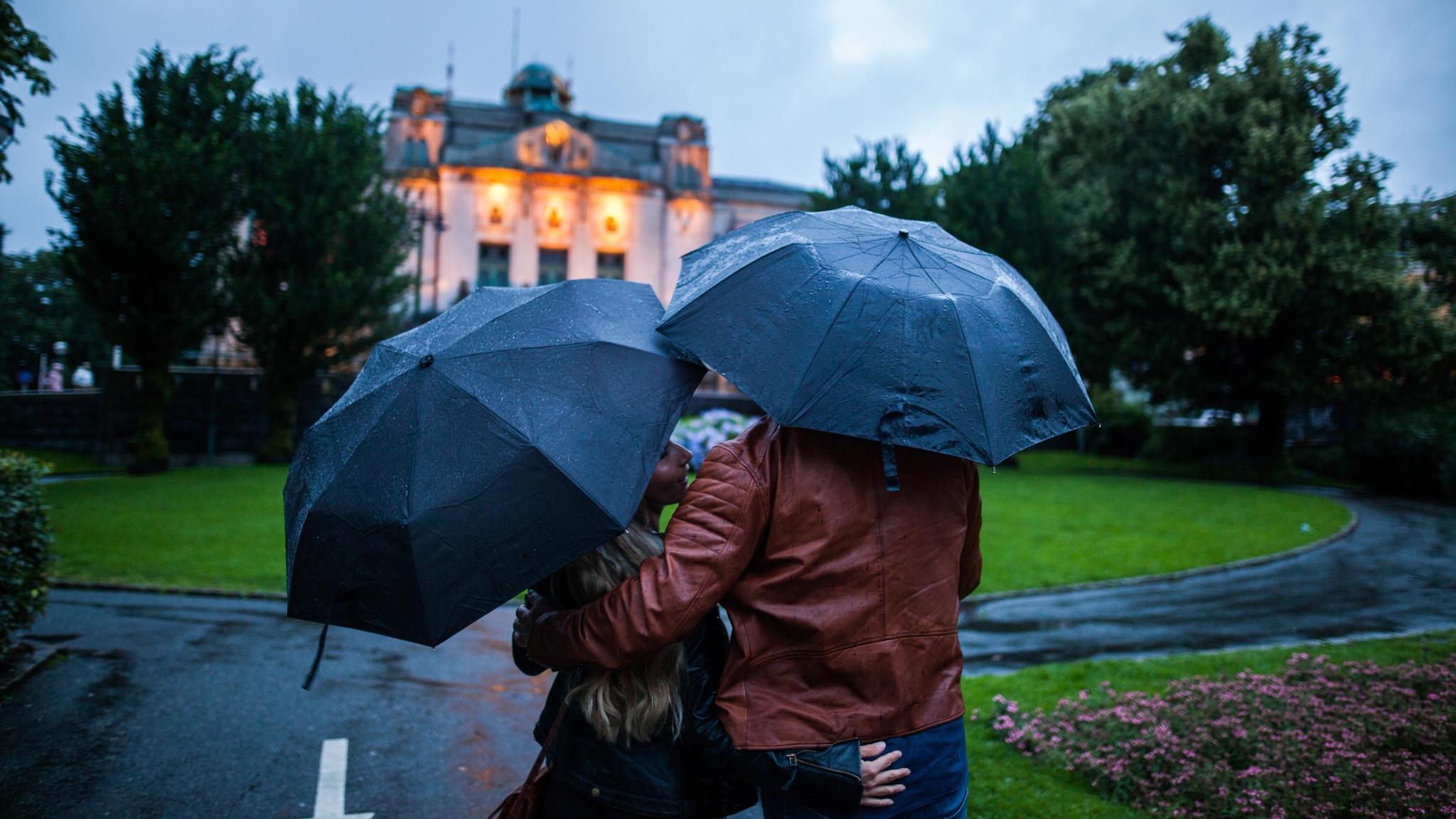 A couple, each with their own umbrella, holding around each other in the rain in Bergen, Fjord Norway. In the background, there is an official looking building.