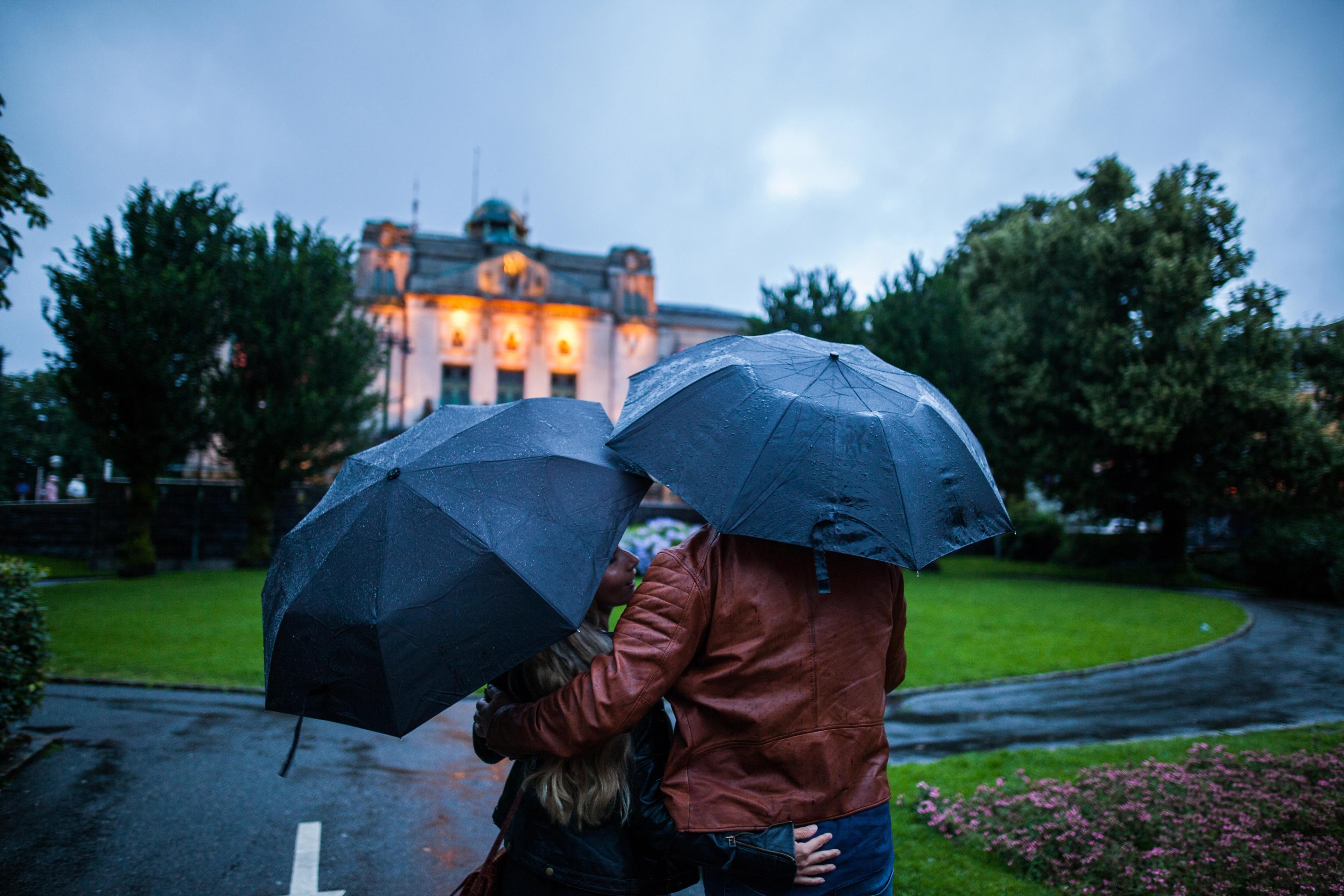 A couple, each with their own umbrella, holding around each other in the rain in Bergen, Fjord Norway. In the background, there is an official looking building.
