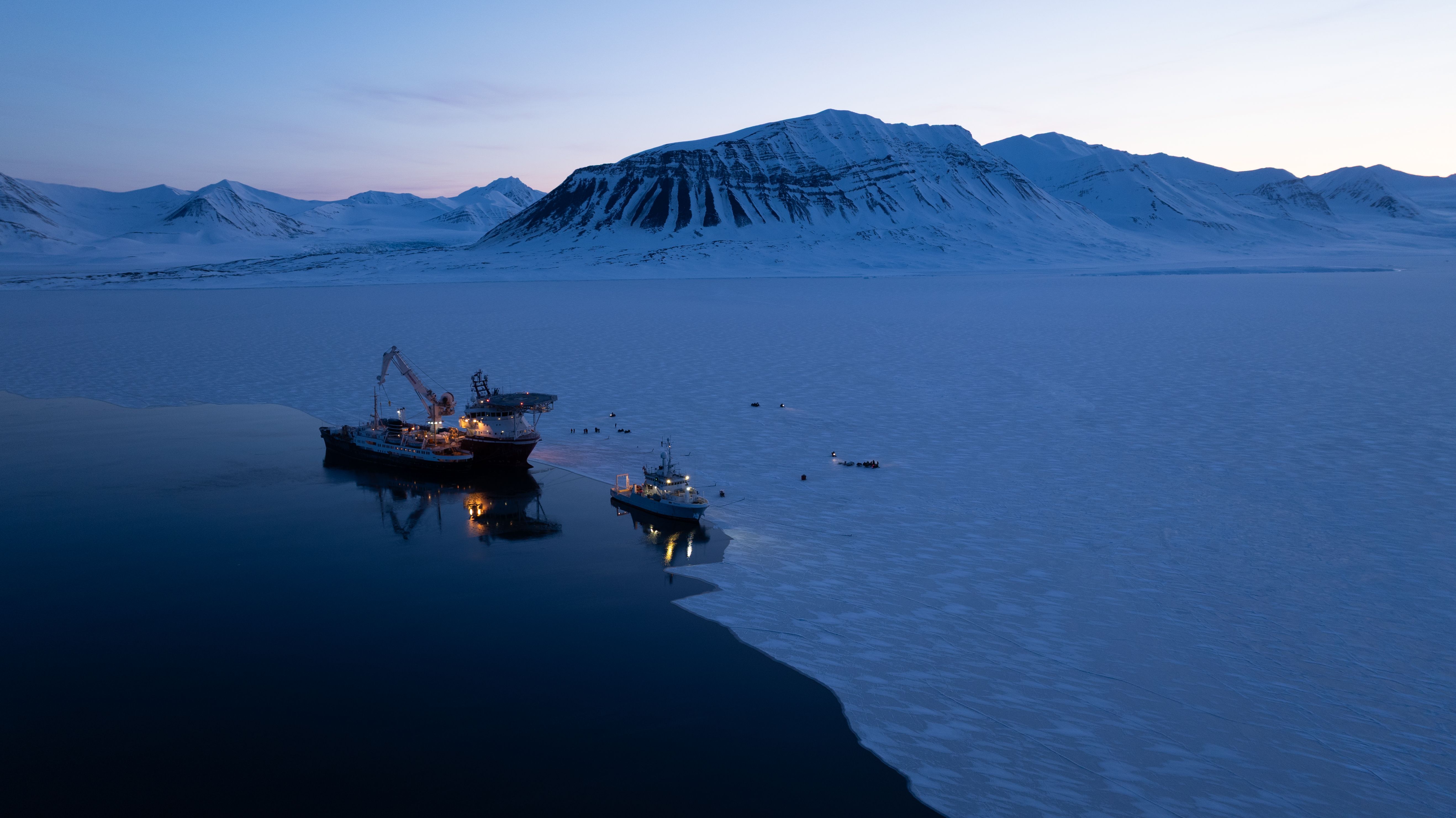 Three ships docked near thick sheet ice below mountains.