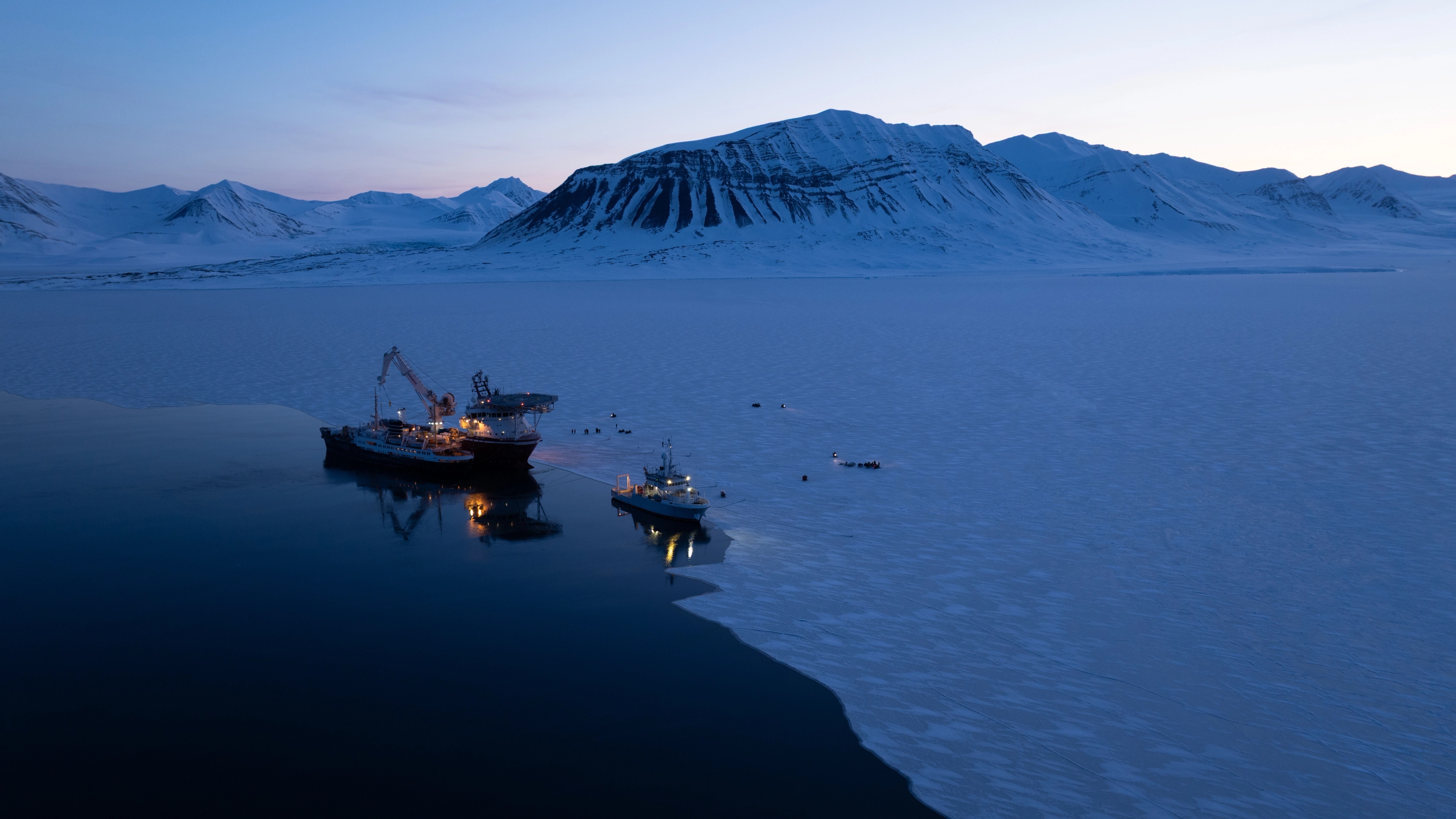 Three ships docked near thick sheet ice below mountains.
