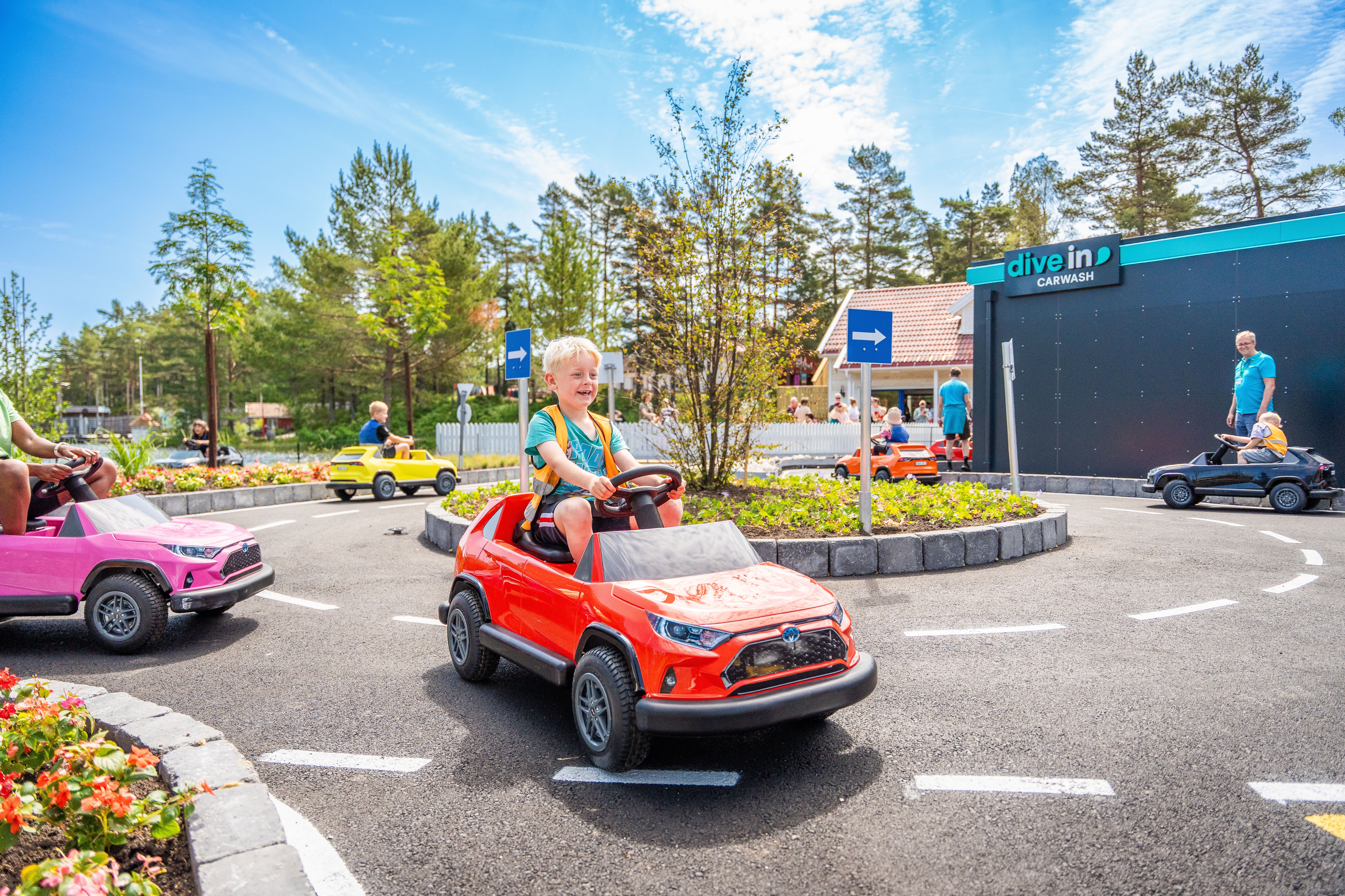Blonde boy driving red toy car around a roundabout at an amusement park.