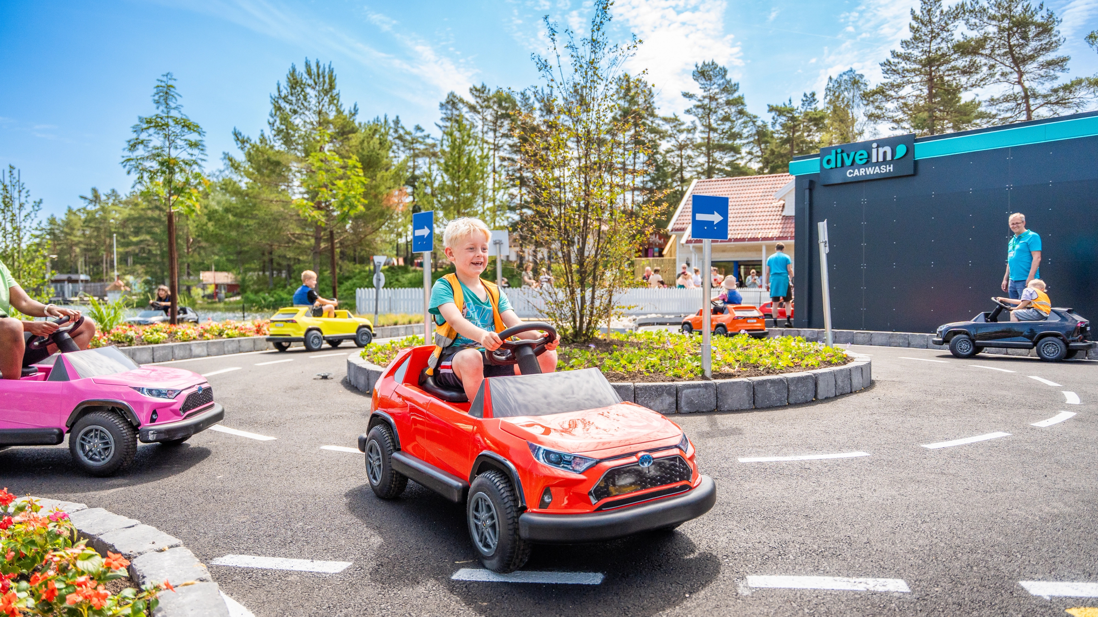 Blonde boy driving red toy car around a roundabout at an amusement park.