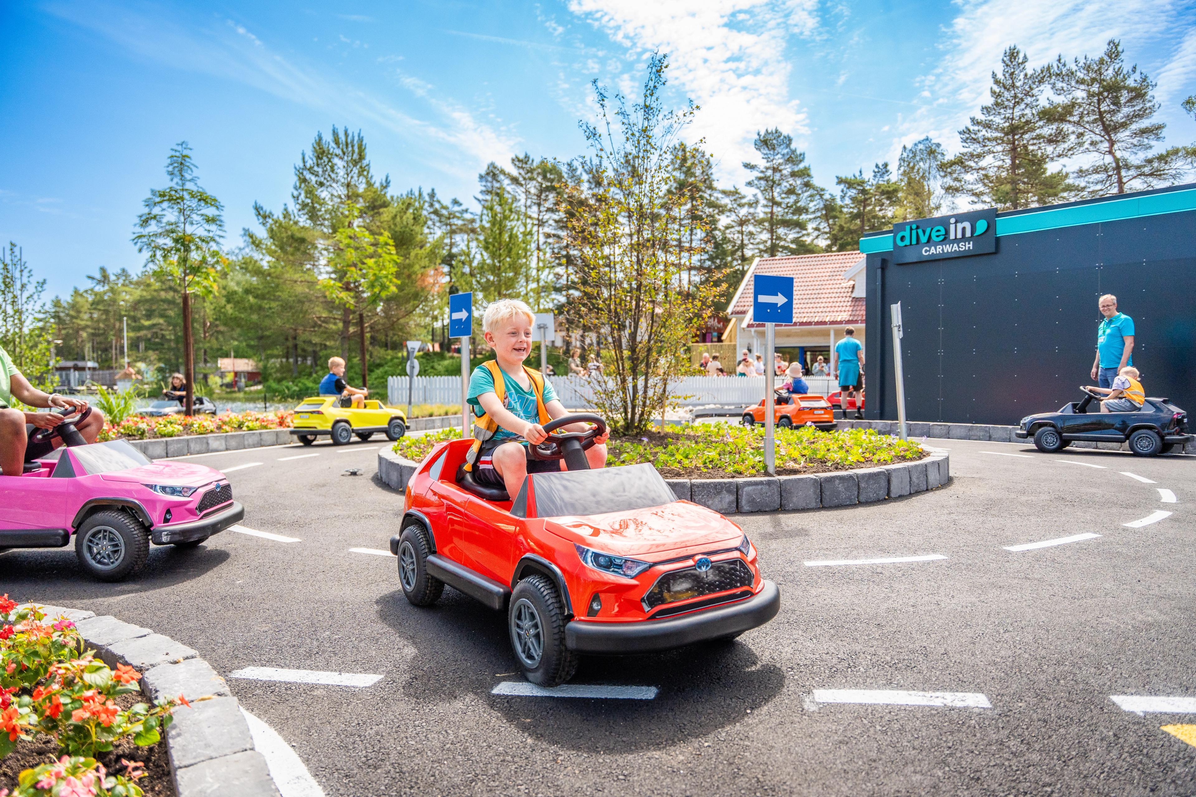 Blonde boy driving red toy car around a roundabout at an amusement park.