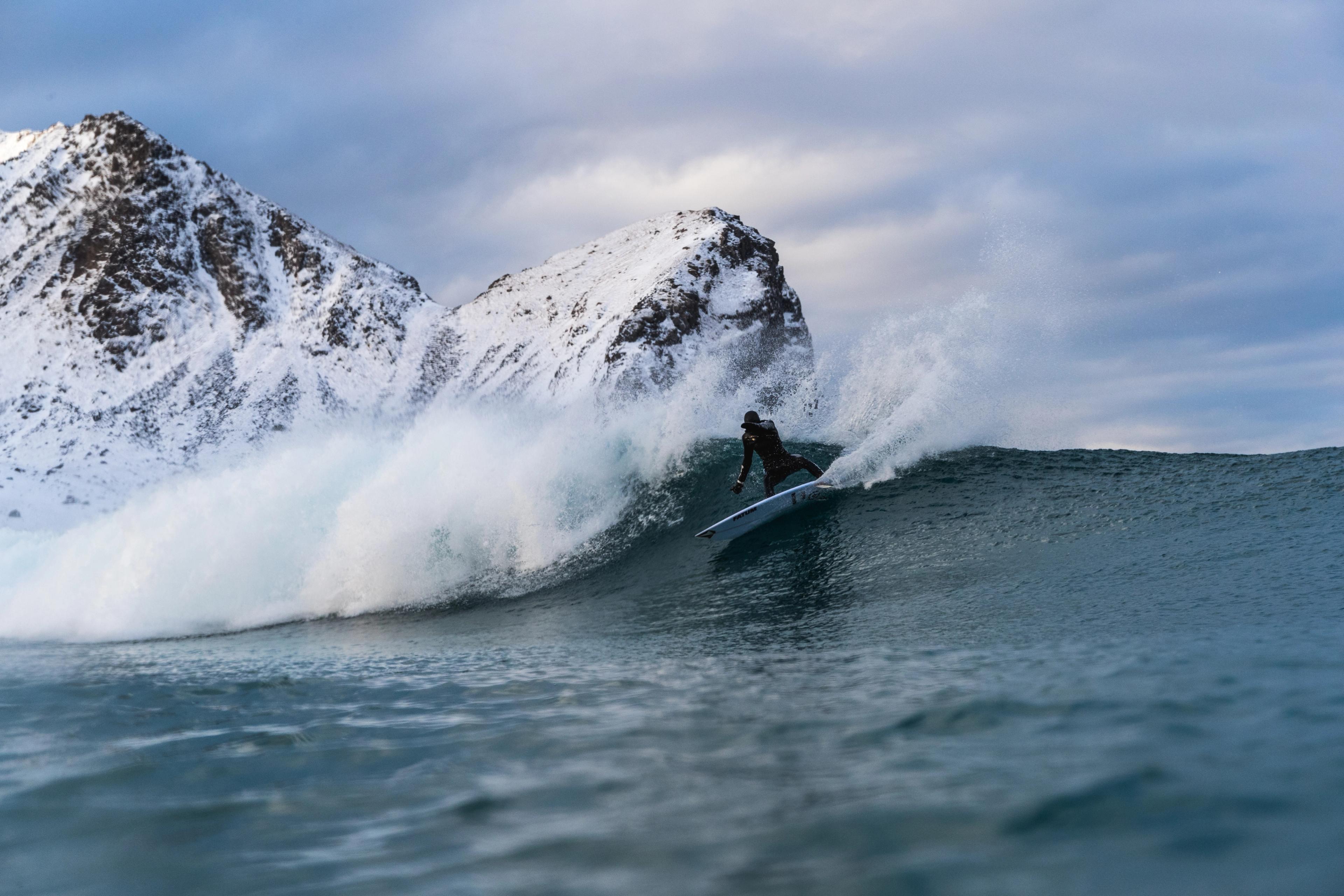 A surfer catches a breaking wave with a snow-covered mountain in the background in Lofoten in Northern Norway